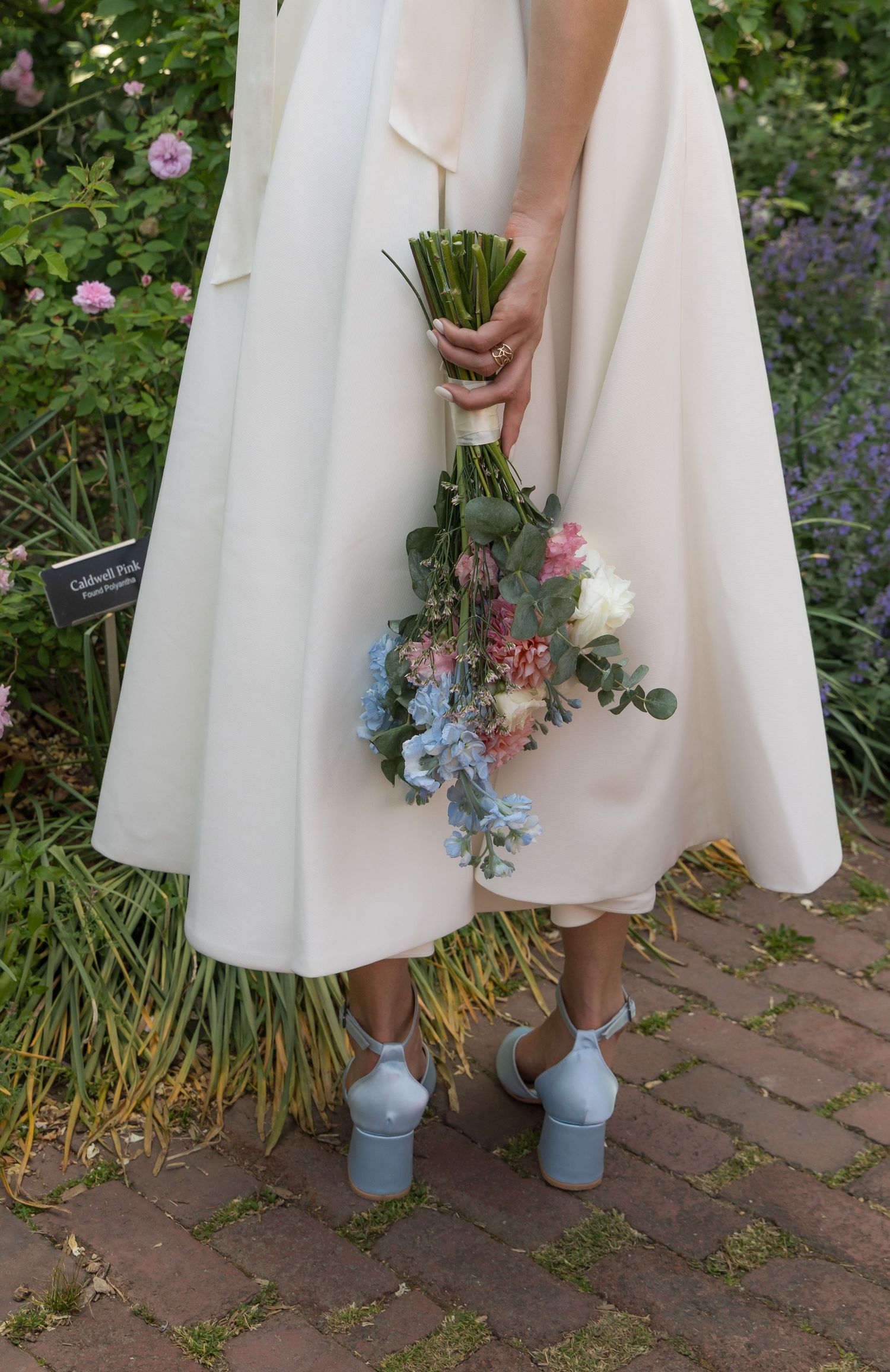 Bride in white dress holds colorful bouquet wearing blue block heel shoes on brick path.