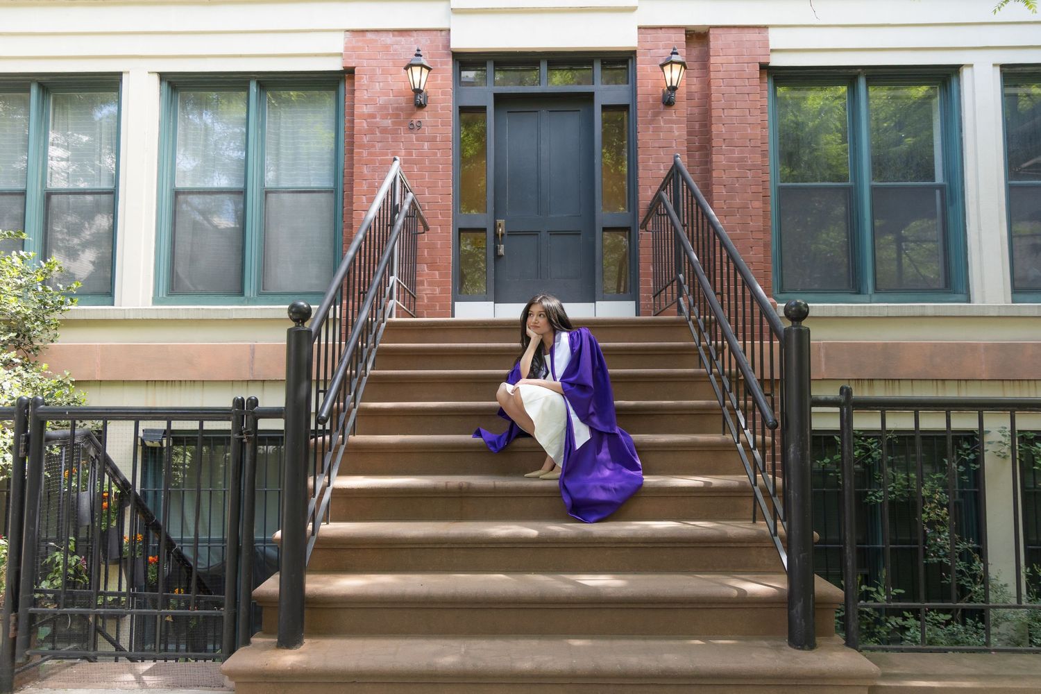 A graduate in purple cap and gown sits on brownstone steps outside a brick building.