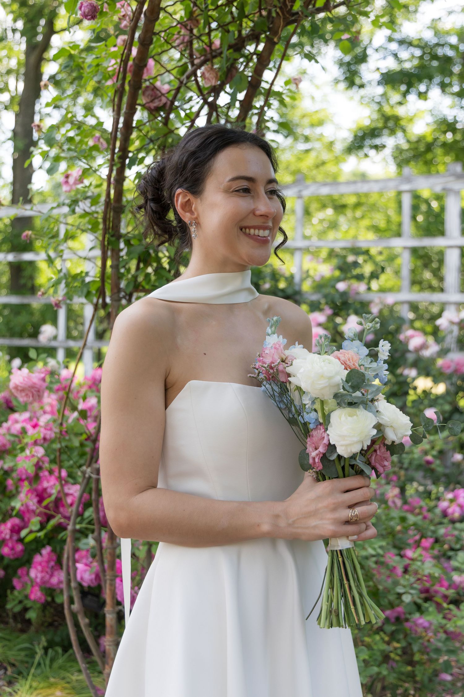 Smiling bride in white strapless gown holding pastel bouquet, standing in lush garden with pink roses.
