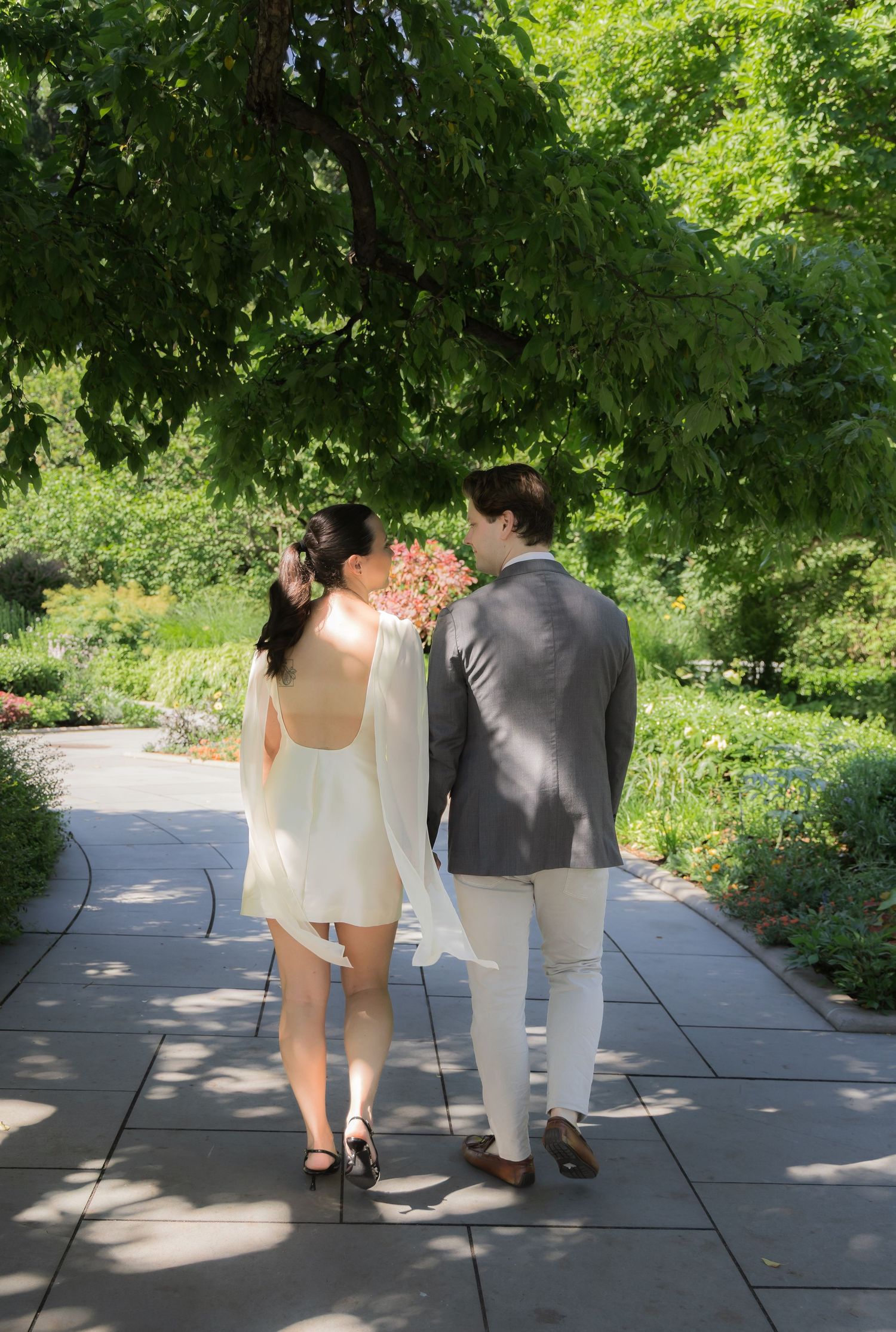 Couple in wedding attire walking hand-in-hand along a garden path surrounded by lush green trees.