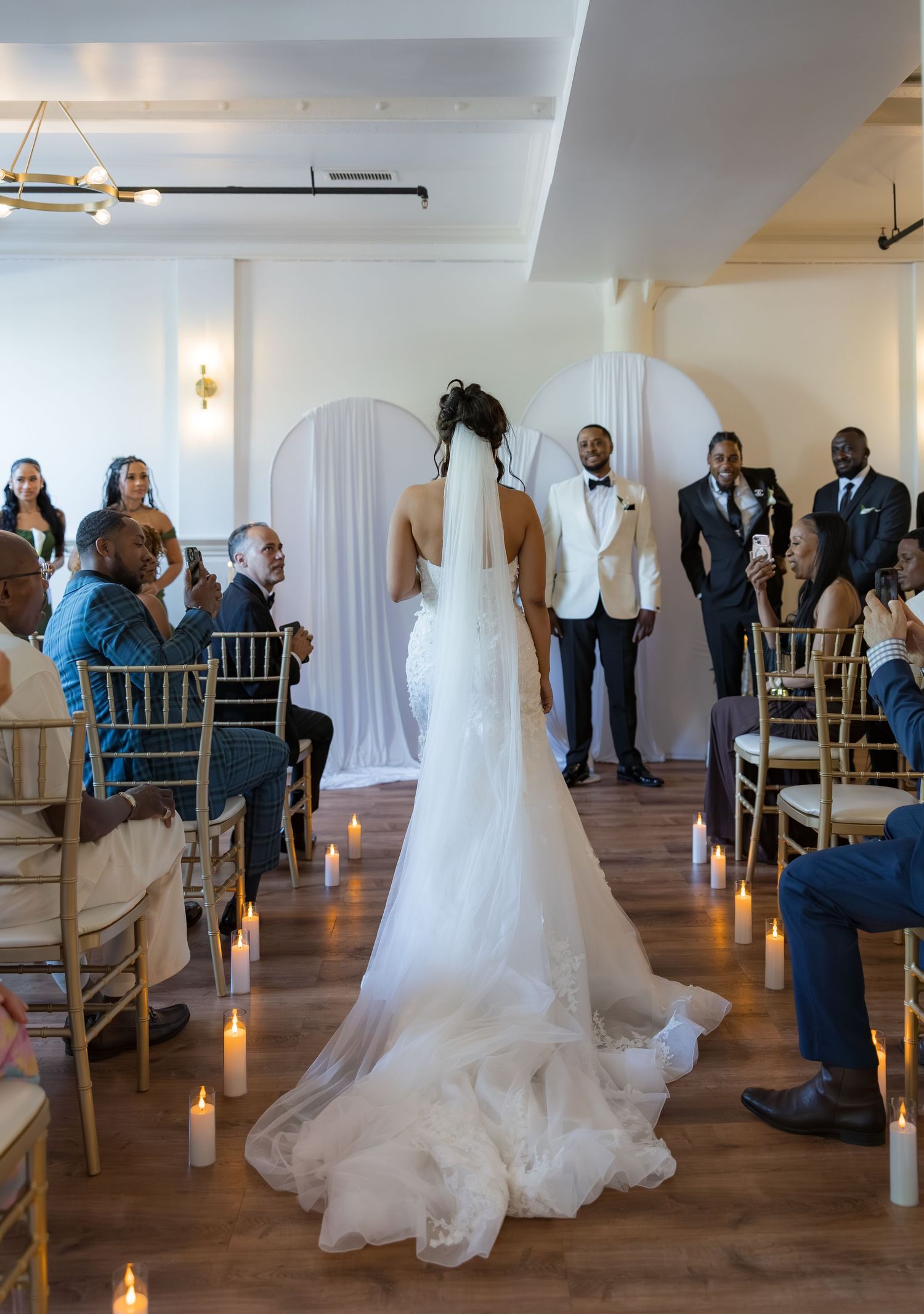 Bride walks down aisle in white gown and veil toward groom, flanked by candles and seated guests in elegant indoor venue.