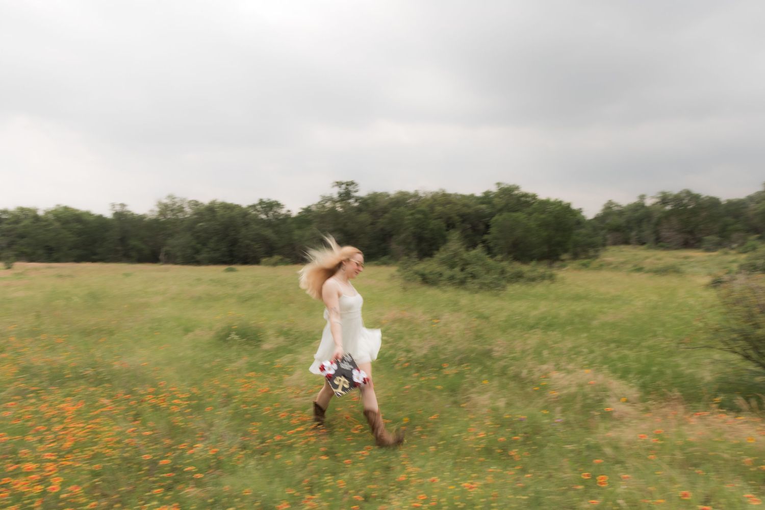 Blurred action photo of a blonde woman in white dress and cowboy boots running in a meadow.