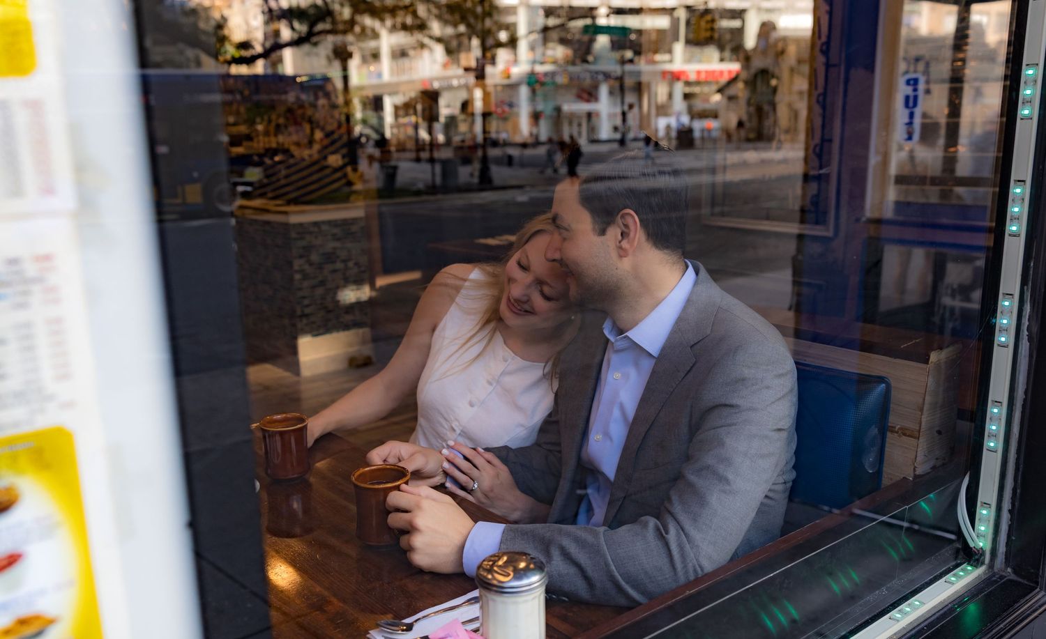 Couple sharing a romantic moment at a restaurant window table, smiling and holding hands on a city street backdrop.