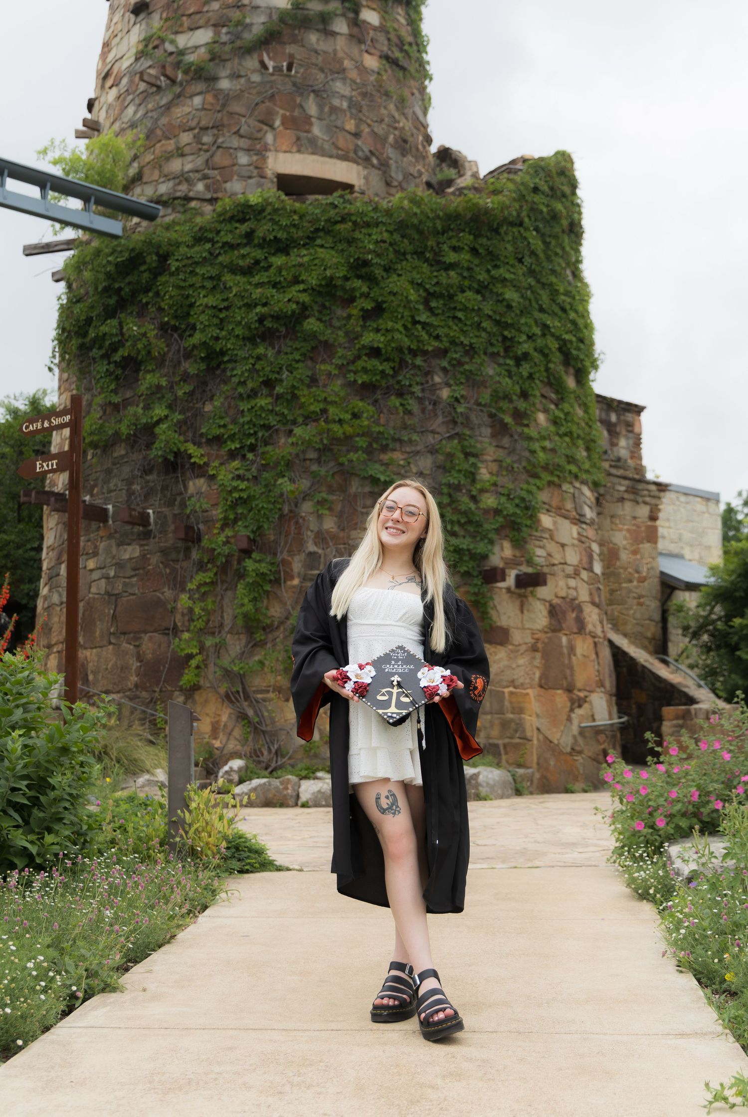 Smiling blonde graduate in black gown holding decorated cap outside stone castle tower.