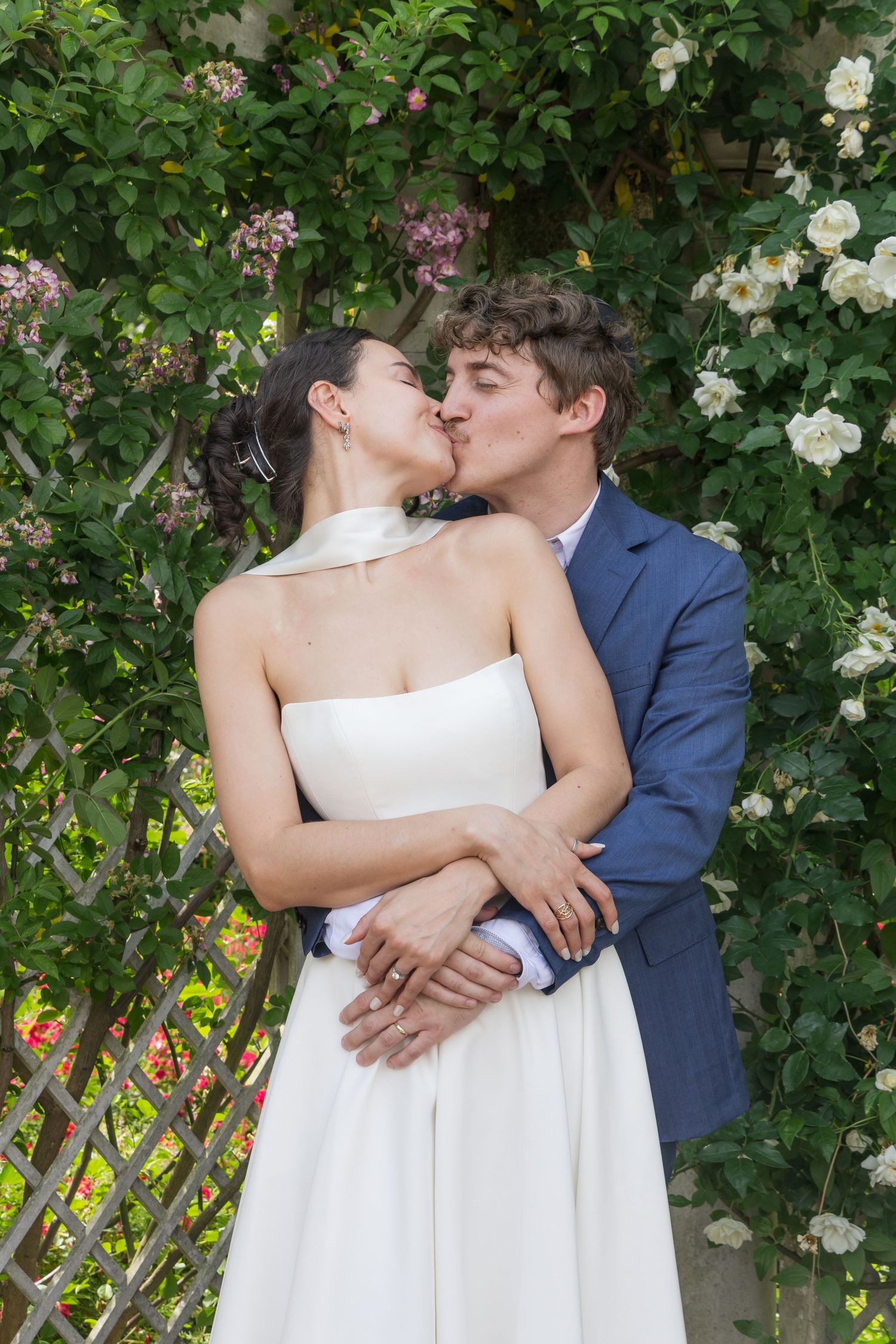 A bride and groom share a romantic kiss in a lush garden, surrounded by blooming white and pink roses.