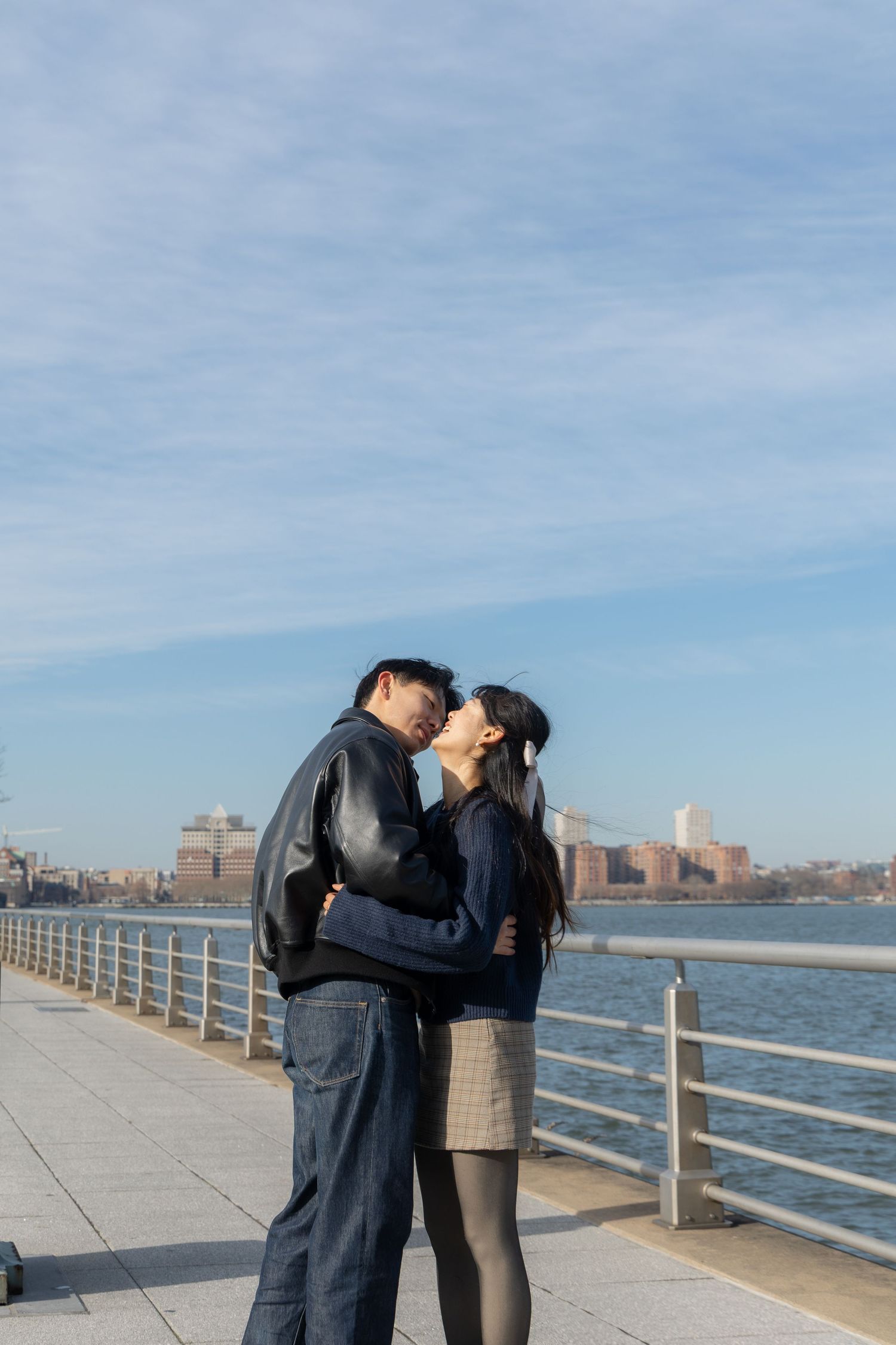 A couple embraces on a waterfront promenade with city buildings visible across the water on a sunny day.