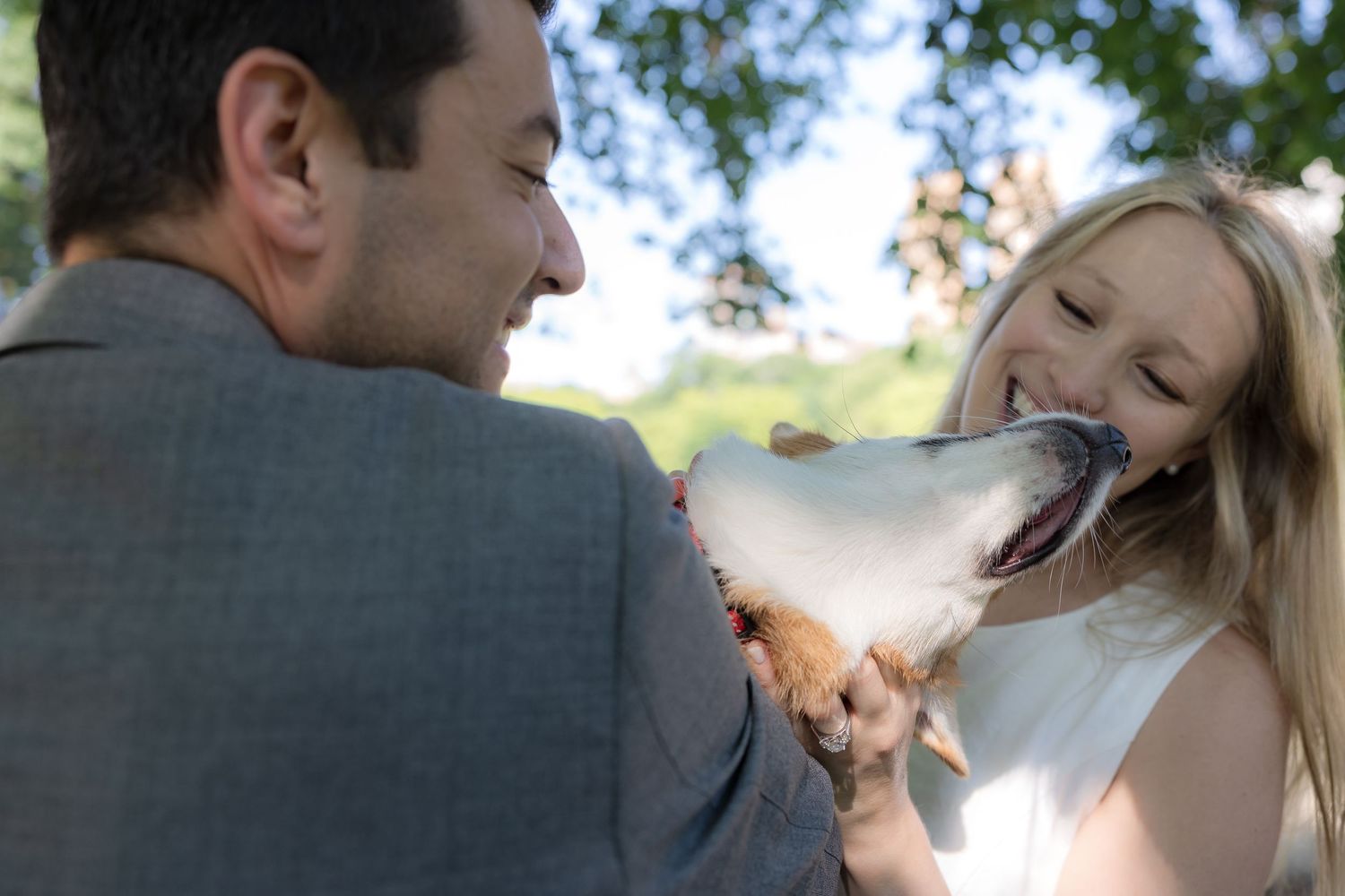 Joyful woman in white dress laughing as a dog playfully licks her face outdoors.