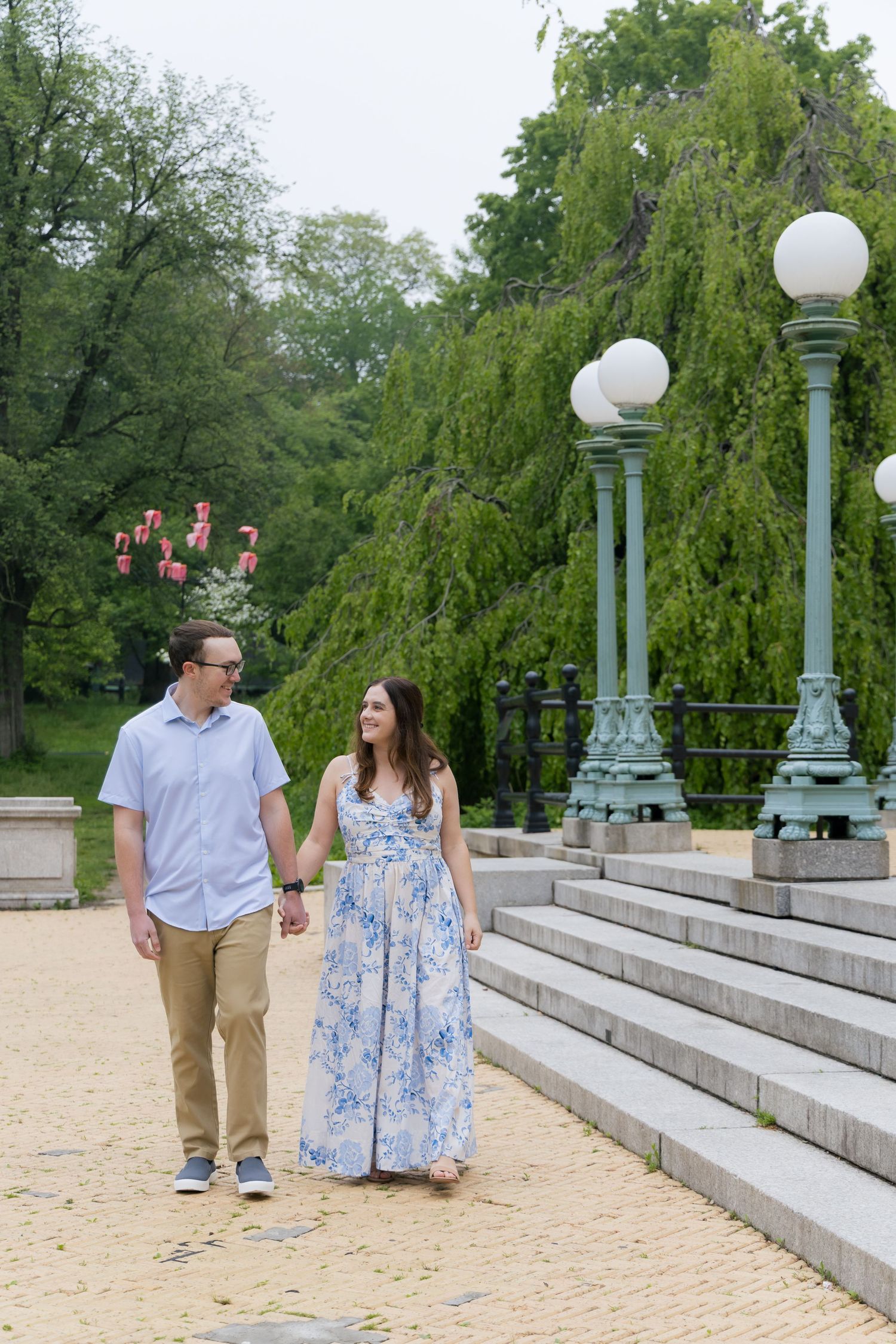 A couple holds hands and walks along a scenic park path with ornate green lamp posts and lush trees in the background.