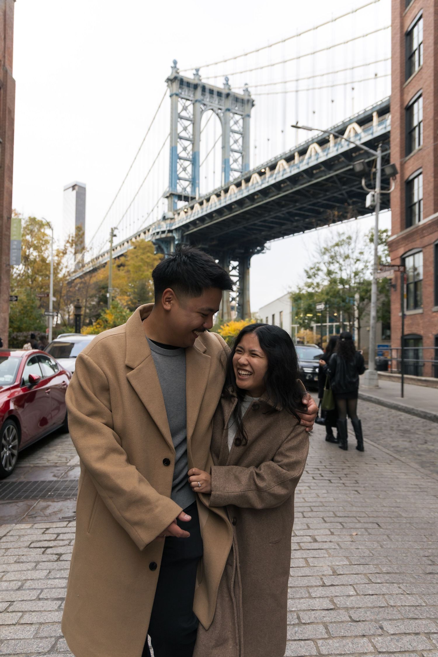 Couple sharing joyful moments in front of Manhattan Bridge on a cobblestone street in DUMBO, Brooklyn.