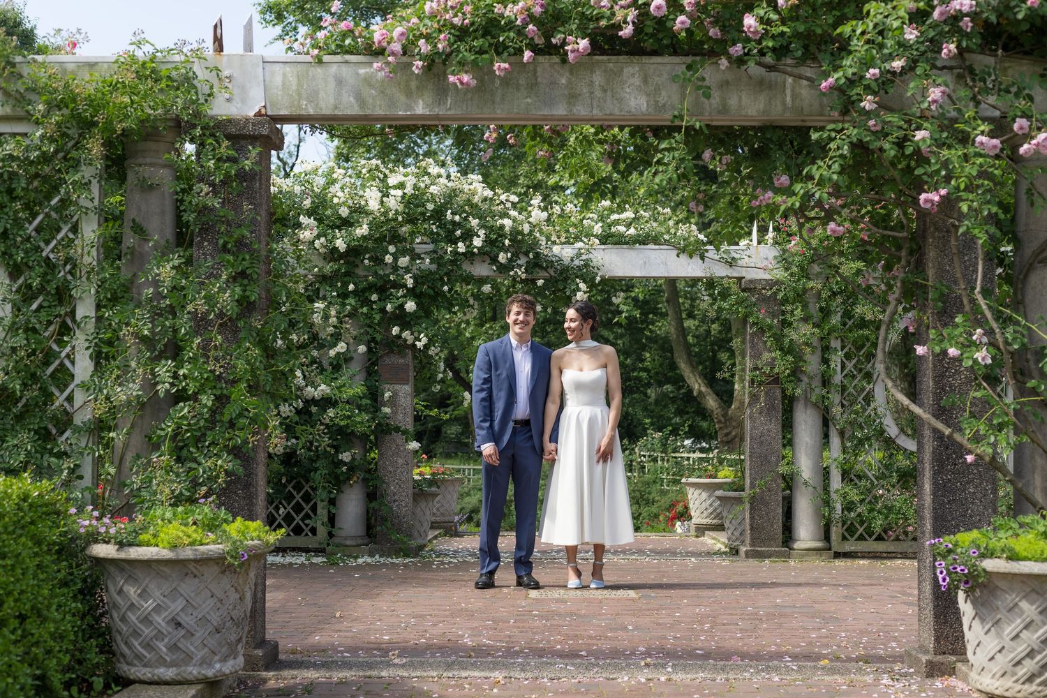 Couple standing under a flower-covered pergola in a lush garden, man in blue suit and woman in white dress.