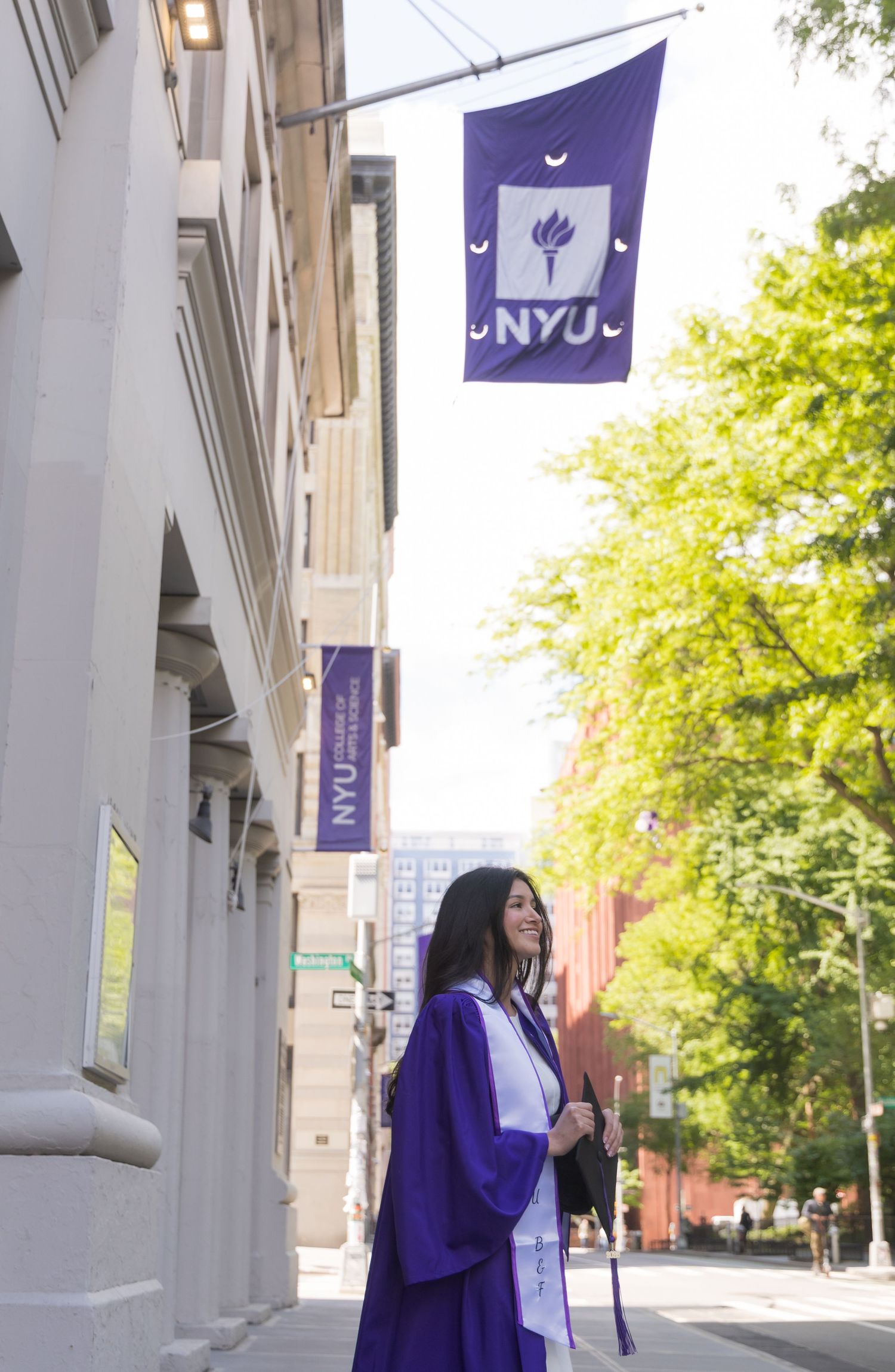 NYU graduate in purple cap and gown smiling under NYU flag on sunny campus street in New York City.