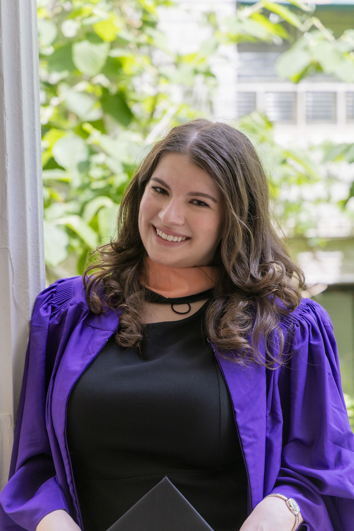 Smiling young woman in purple graduation gown holding diploma, posing outdoors with green foliage background.