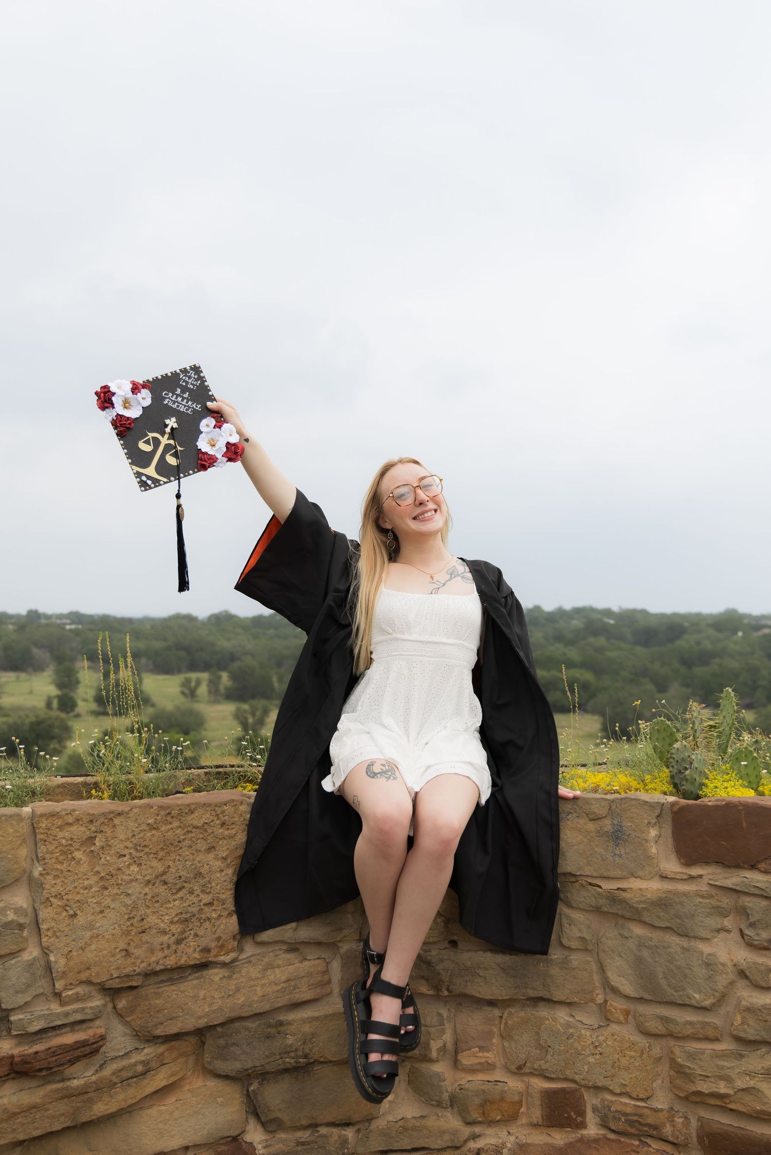 Joyful blonde graduate in black gown raising decorated mortarboard on stone wall outdoors.