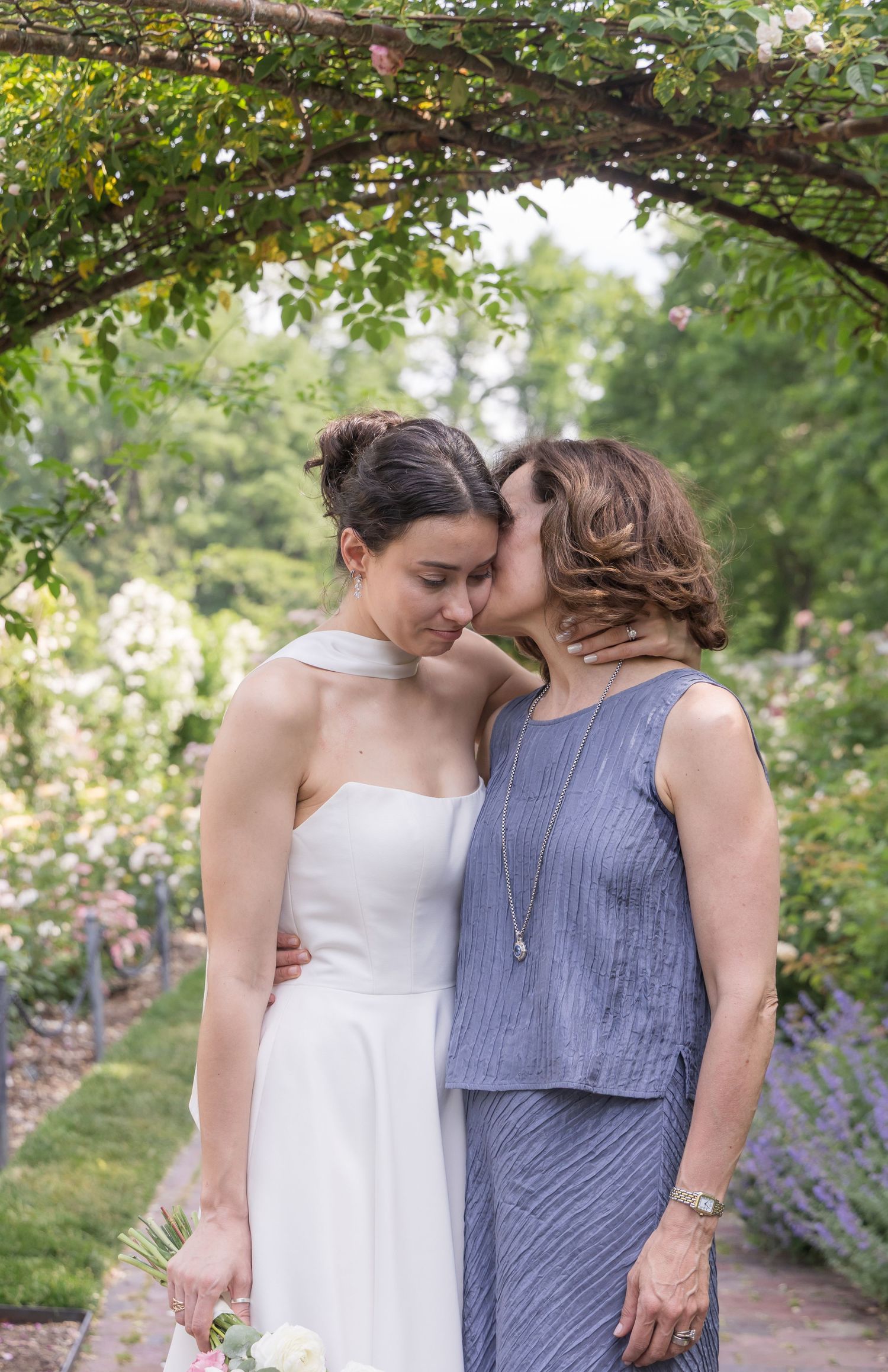 Bride in white dress receives a kiss on the cheek from an older woman in a blue outfit in a lush garden setting.