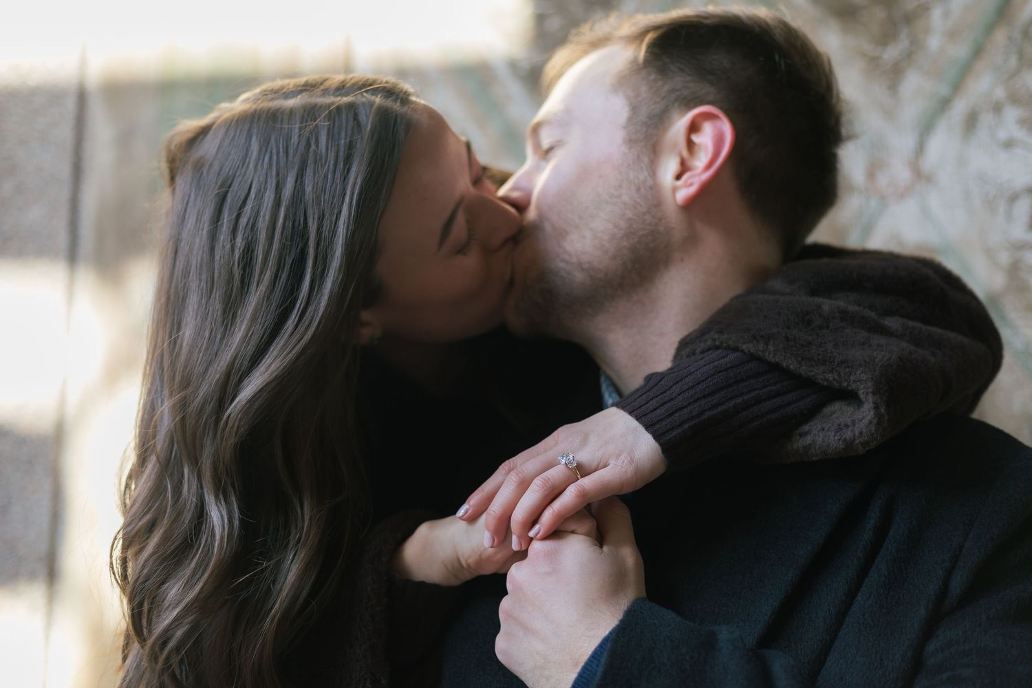 A close up of a couple kissing under the archway of Bethesda Terrace with a close up of her engagement ring.