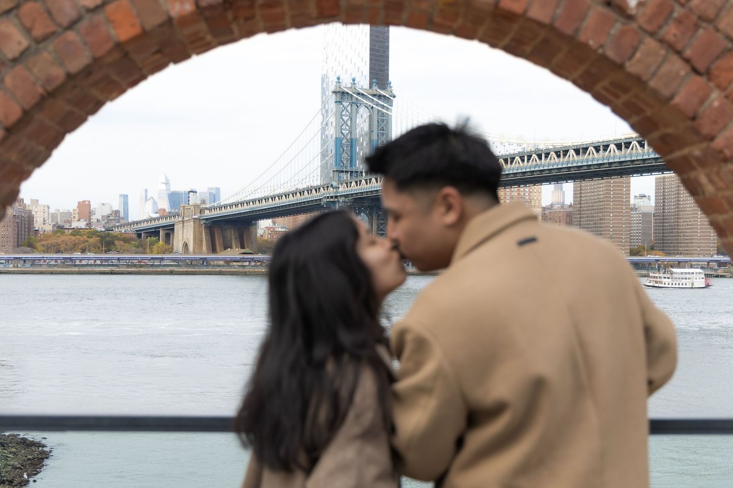 A couple shares a romantic moment along the waterfront with a bridge visible in the background.