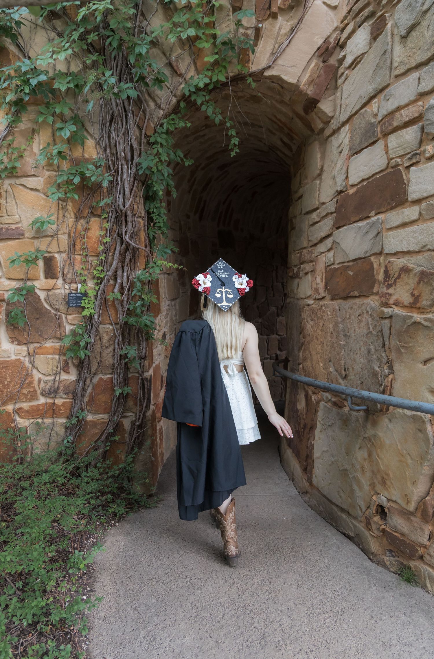 Graduate in cap and gown walking through a stone tunnel archway covered in ivy.