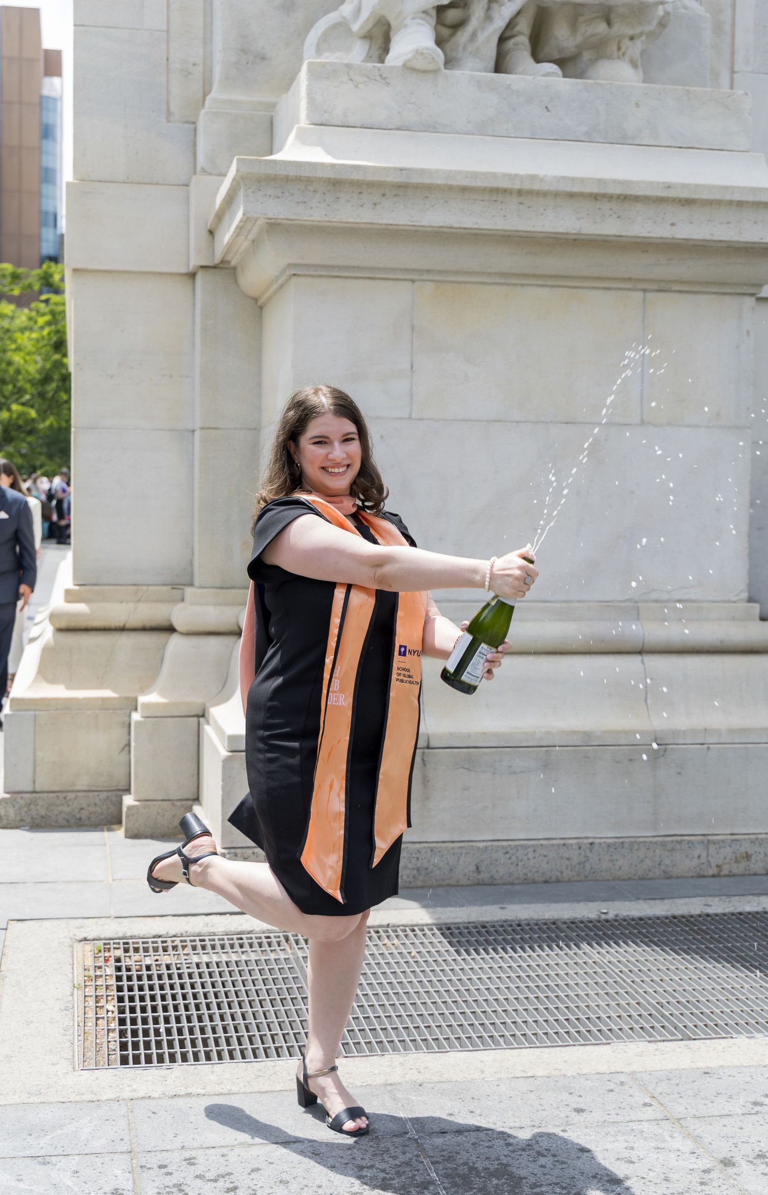 Graduate in black dress and orange sash joyfully sprays champagne bottle while posing on one leg at outdoor ceremony.