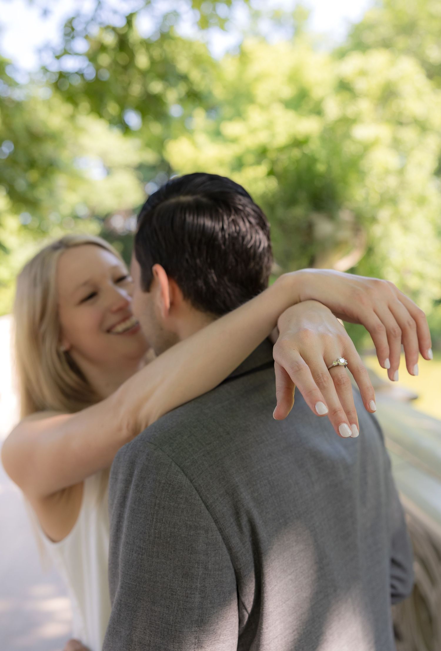 Smiling blonde woman hugging a man outdoors, showing off her engagement ring with green trees in the background.