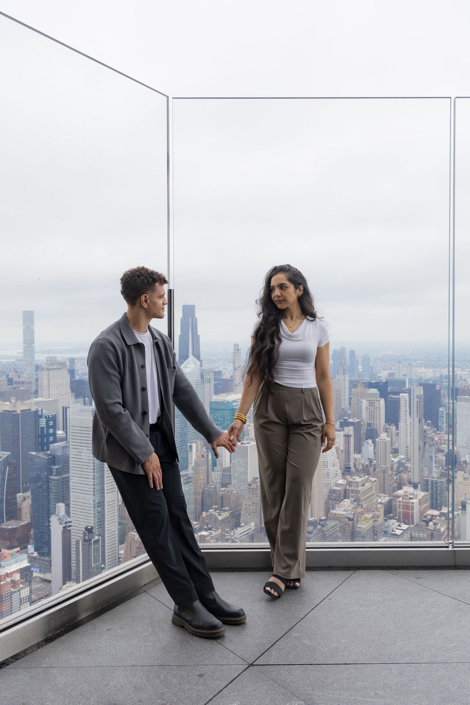 Couple holding hands on a high-rise observation deck overlooking the New York City skyline on a cloudy day.