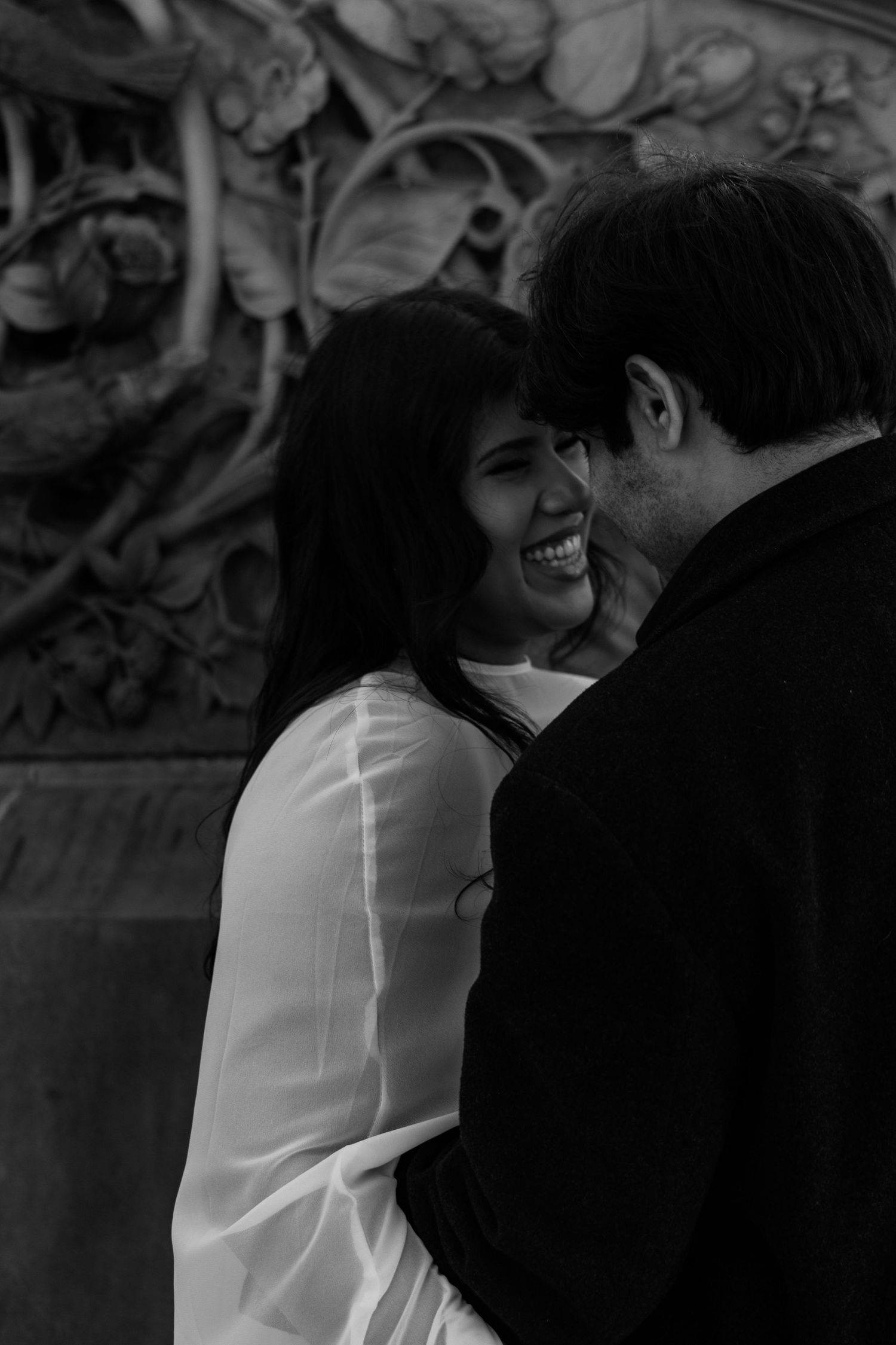 Black and white photo of a smiling woman in white dress sharing a romantic moment with a man outdoors.