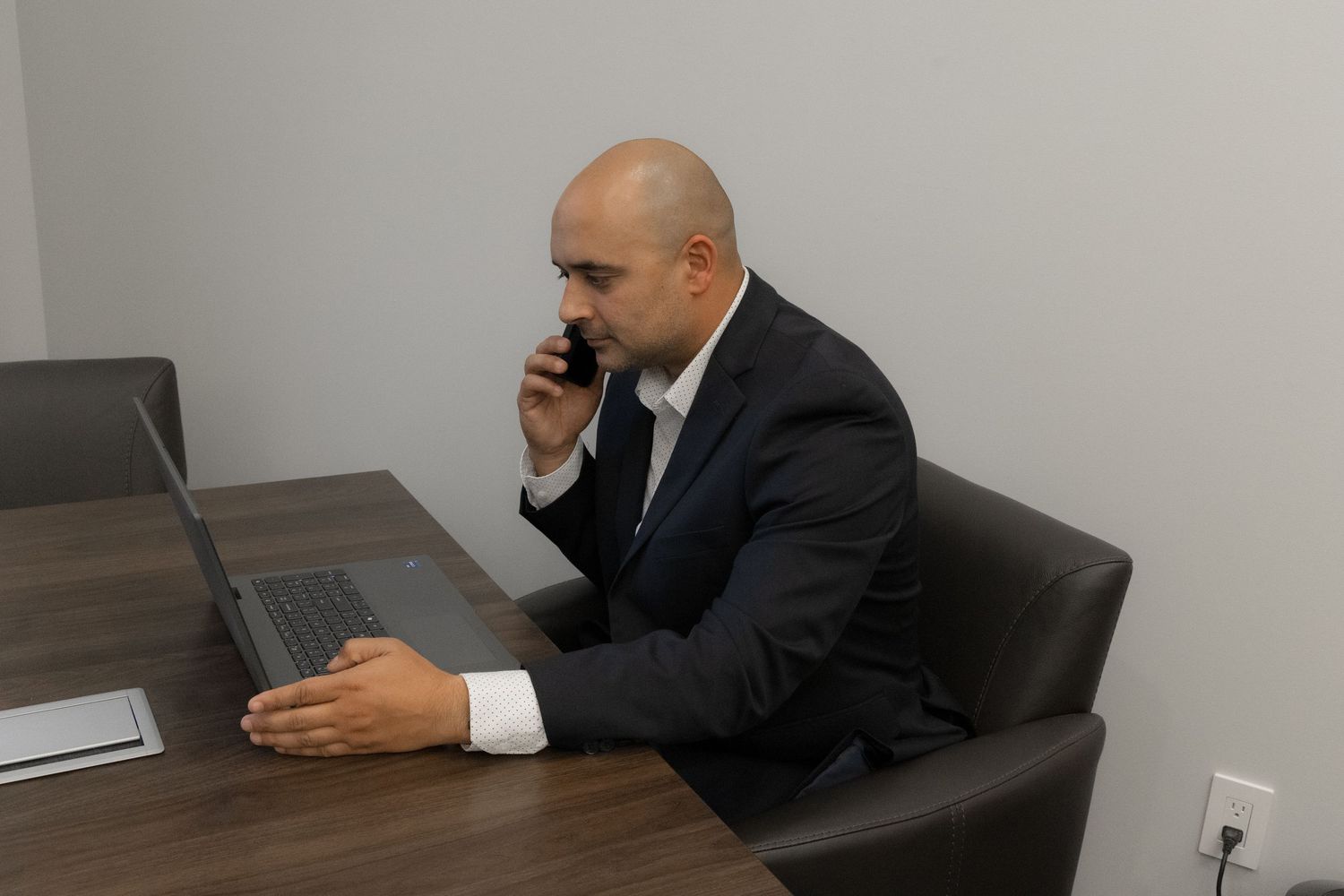 Business professional in dark suit sits at desk looking at laptop while talking on mobile phone.