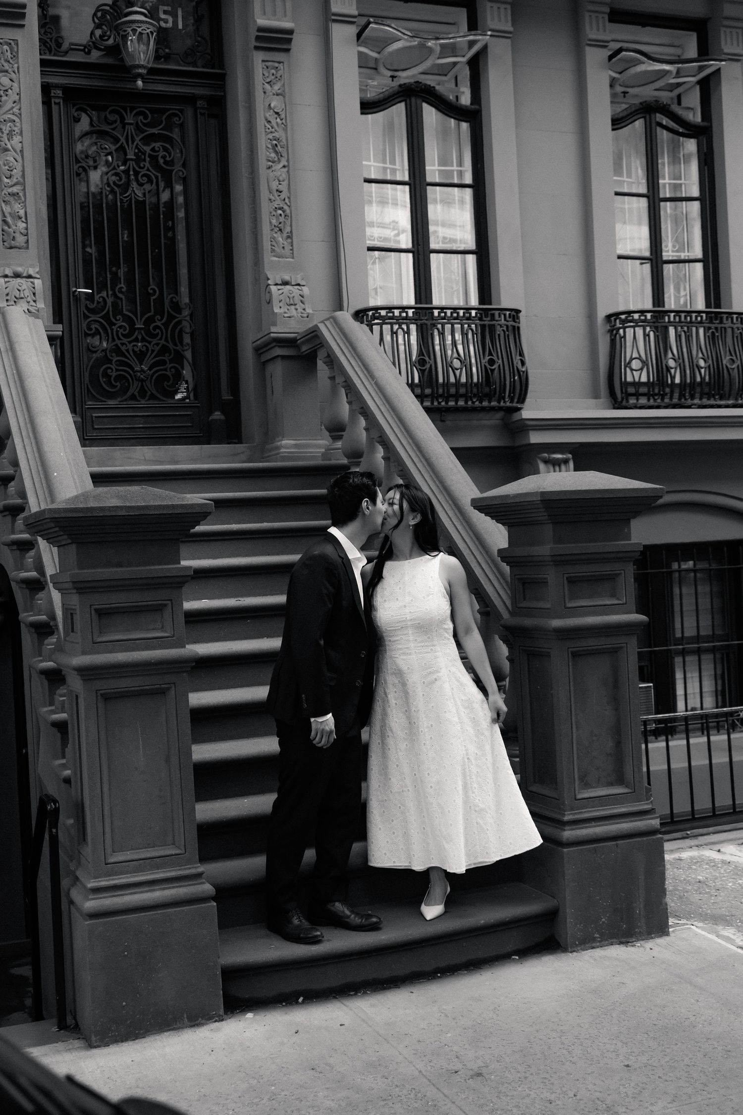 Black and white photo of a couple kissing on brownstone steps, bride in white dress and groom in dark suit.