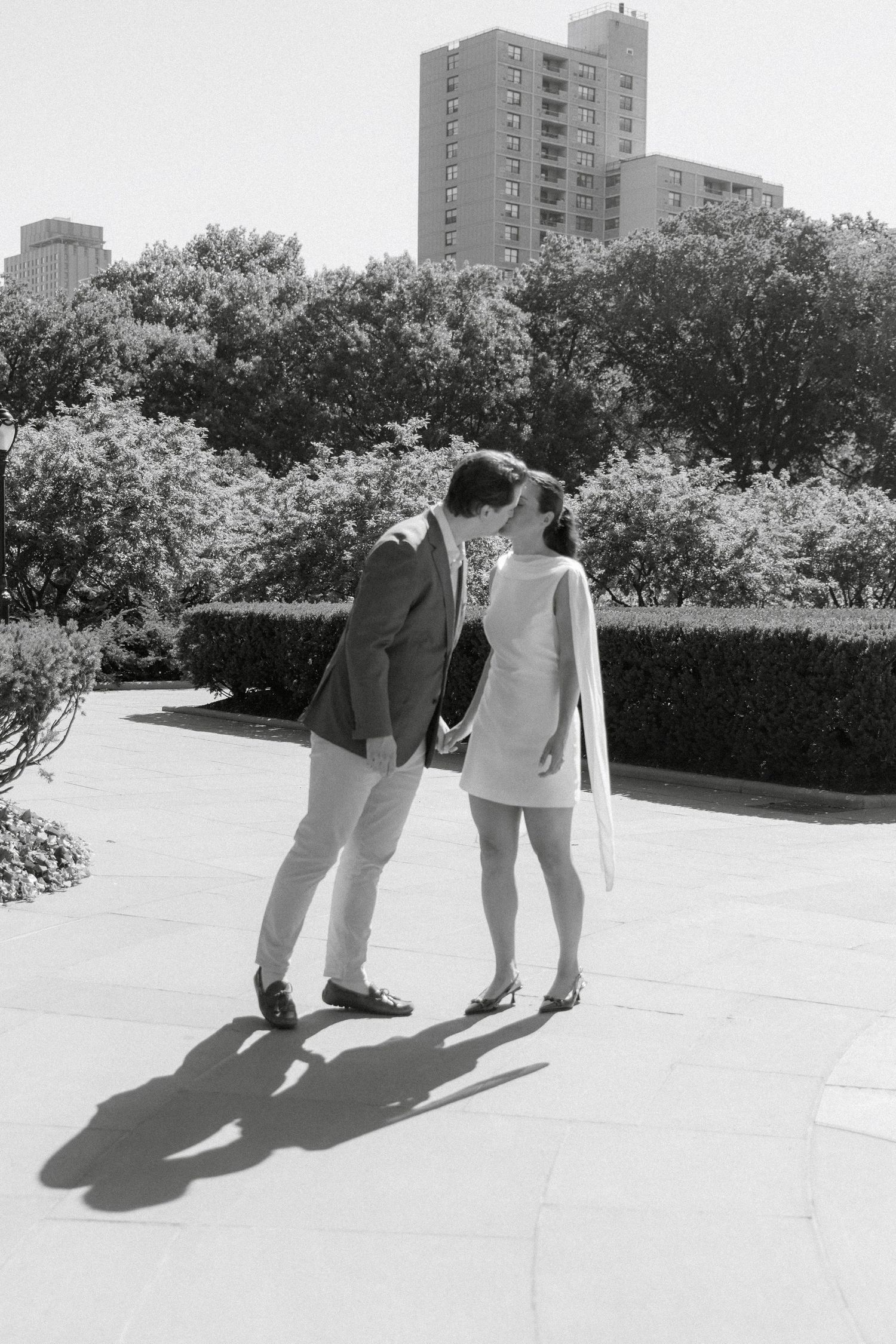Black and white photo of a couple sharing a kiss in a sunny urban park, with trees and a tall building in the background.