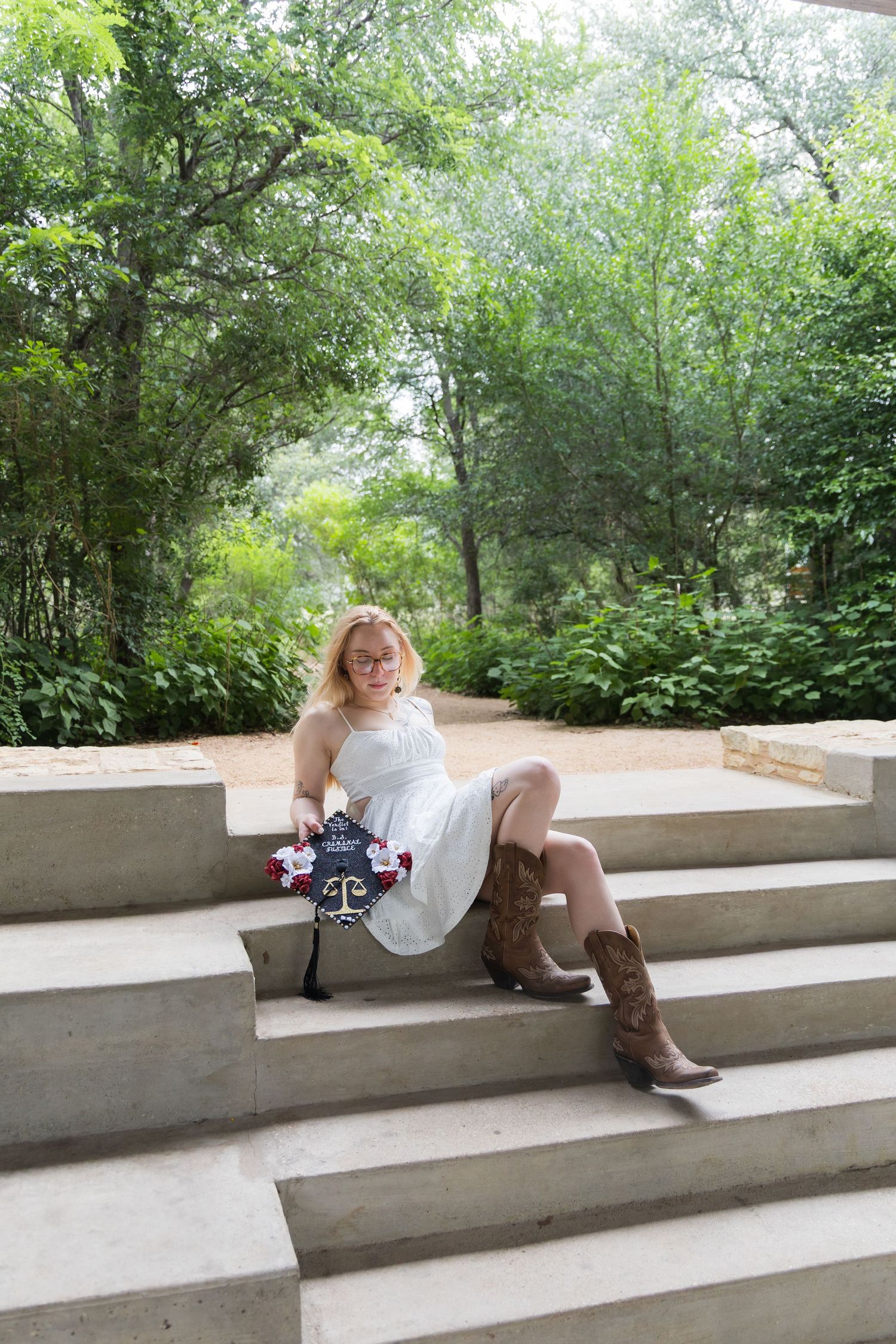 Blonde woman in white dress and cowboy boots sitting on concrete steps holding graduation cap.