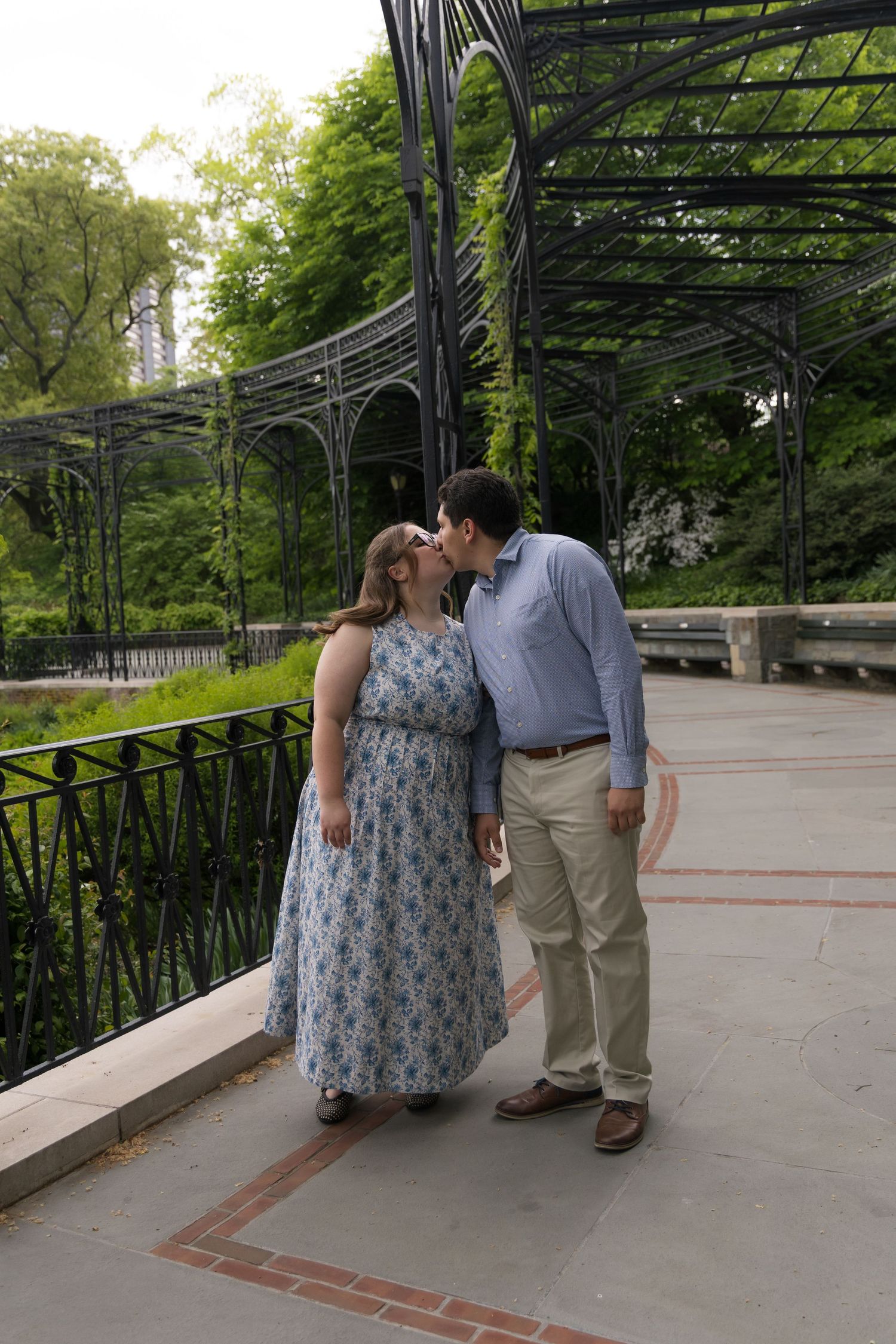 Couple sharing a romantic kiss in a lush park setting under decorative iron pergola on a brick pathway.