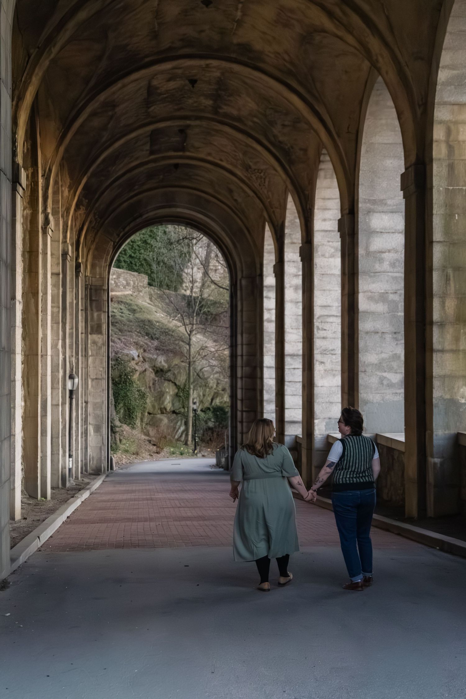 Two women holding hands walk through a grand stone archway corridor with arched columns and a scenic view ahead.