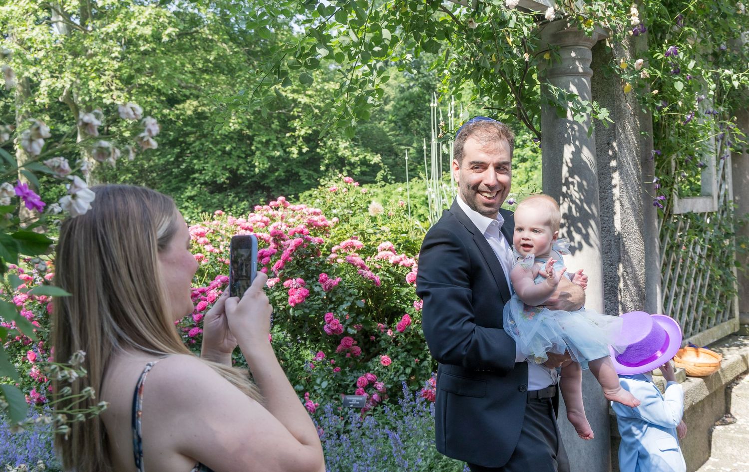 Woman photographing a man in a suit holding a baby outdoors near blooming roses and a pergola.