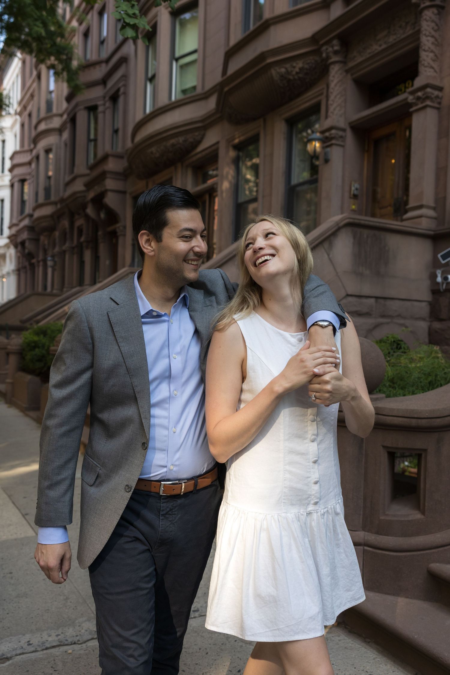 A smiling couple walks arm-in-arm past brownstone buildings on a sunny city street.