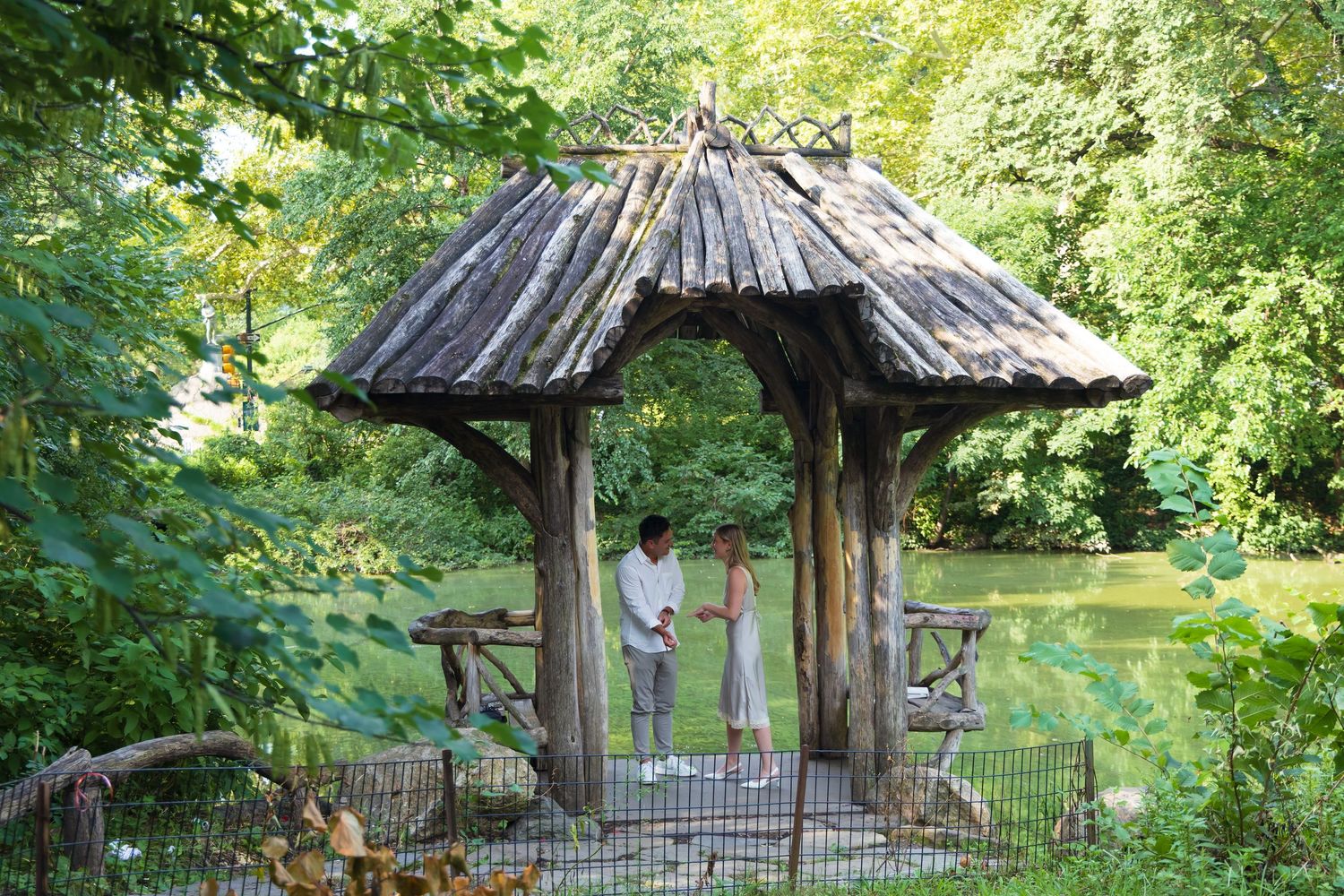 Couple standing under a rustic wooden gazebo surrounded by lush green trees near a tranquil pond.