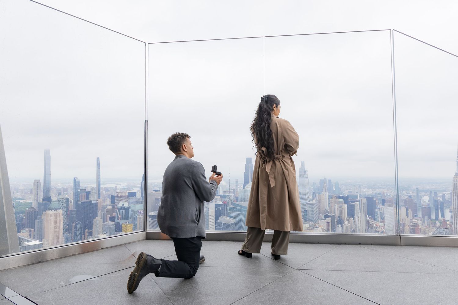 Man proposing on one knee with engagement ring at NYC skyline observation deck overlook.