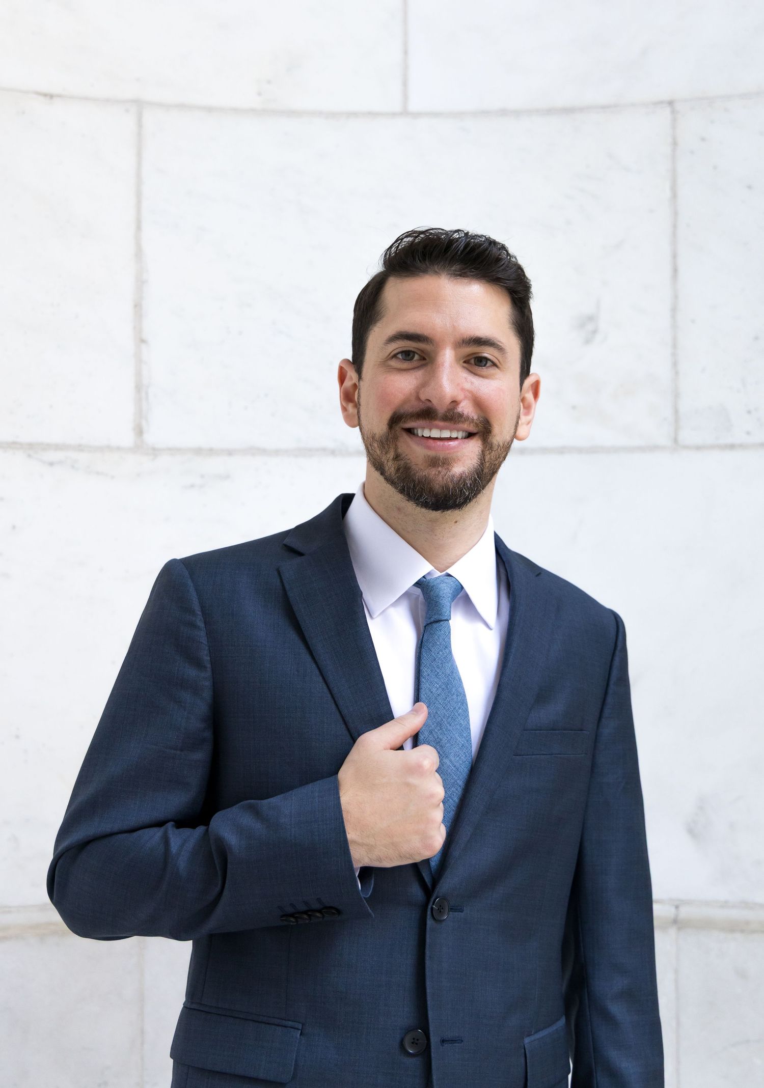 Professional wearing a navy suit and blue tie adjusts his collar against a white brick wall background.