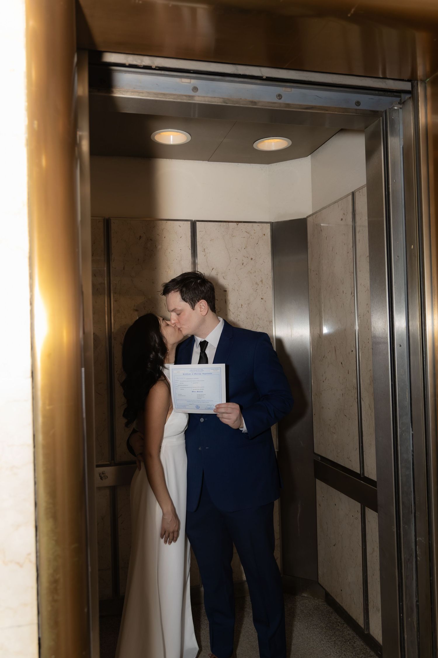 Newlywed couple sharing a kiss in a gold elevator, groom in navy suit holding marriage certificate.