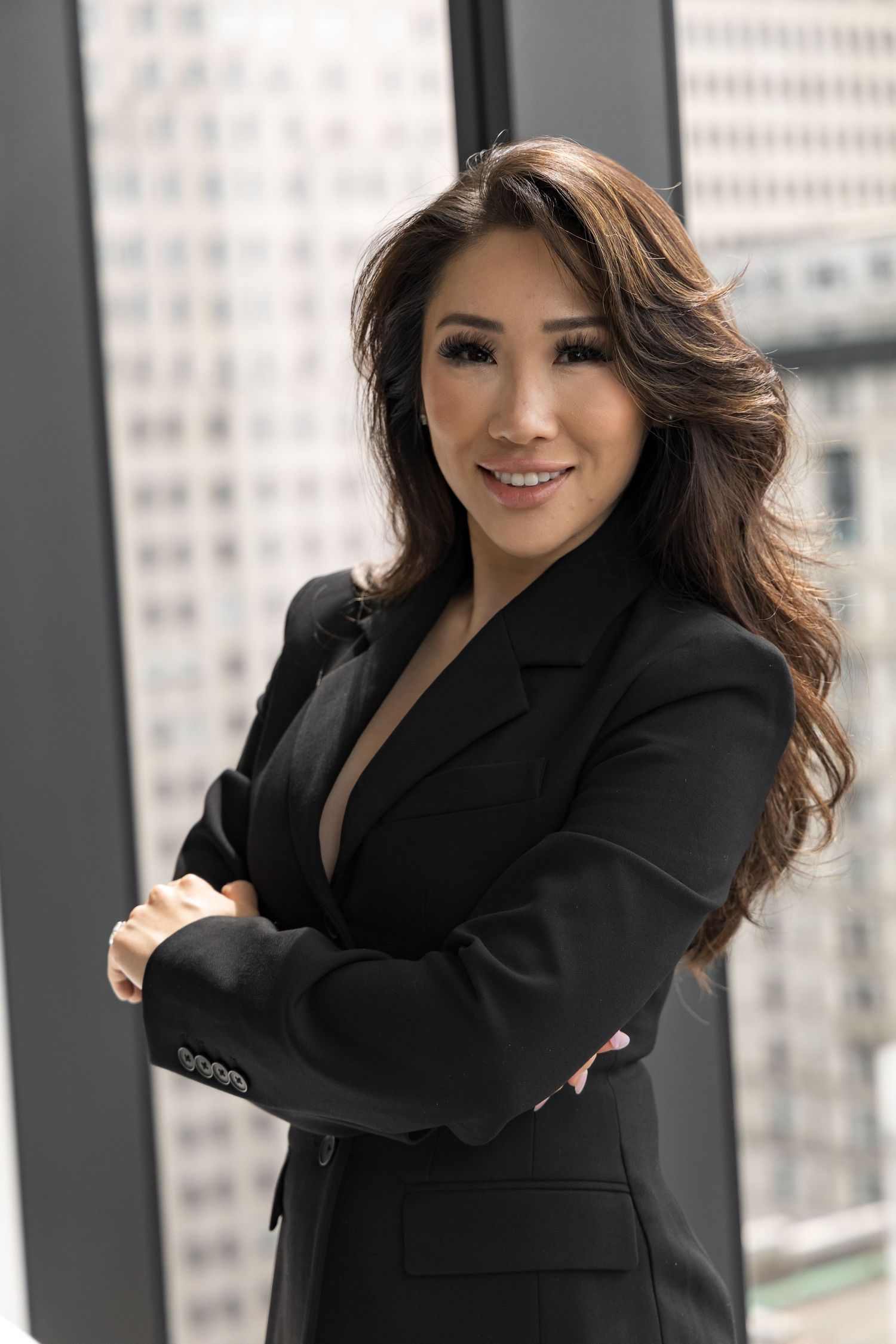 Professional woman in black blazer poses confidently against urban office window background.