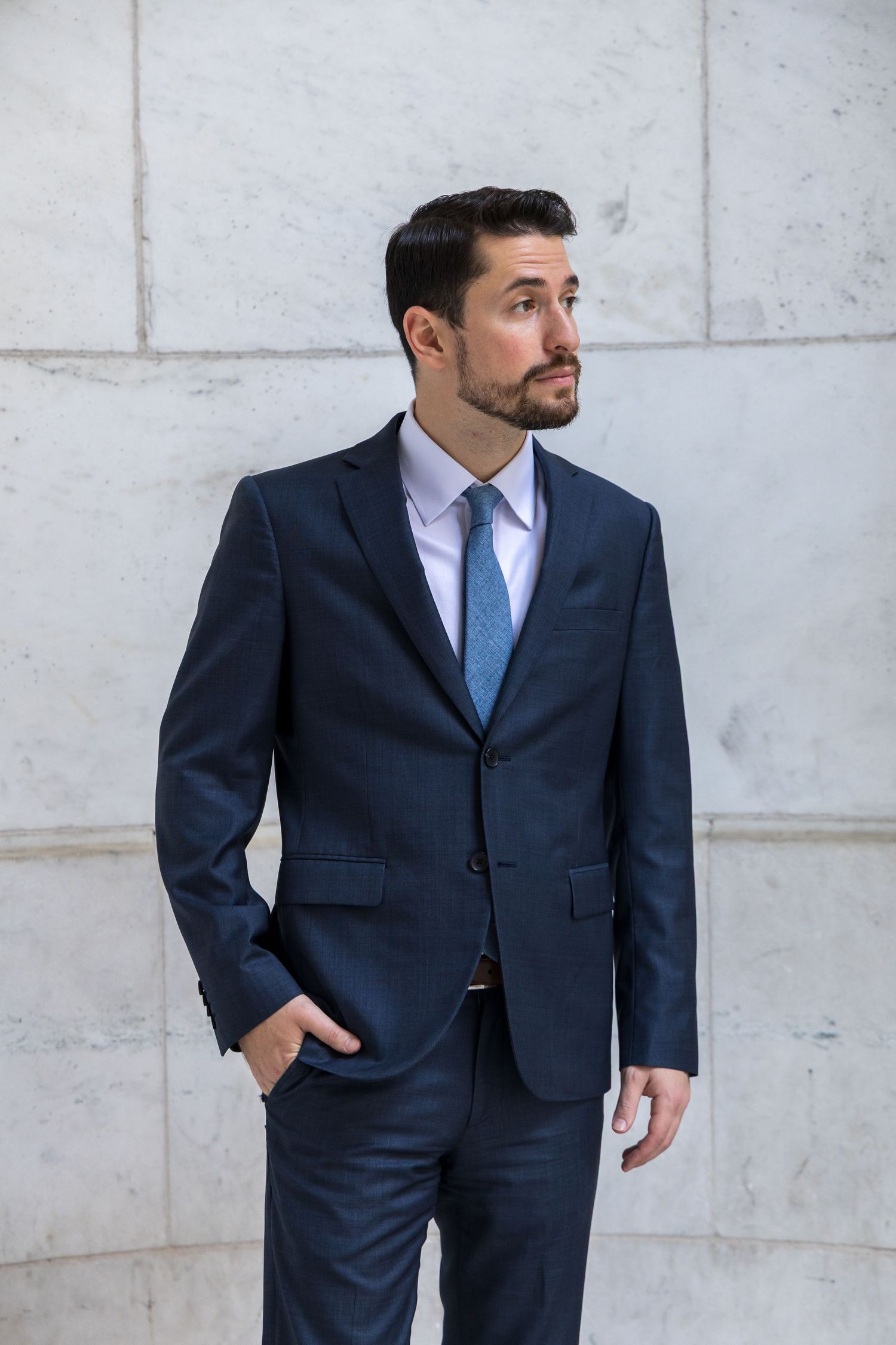 Professional wearing a navy business suit poses against a light concrete wall background.