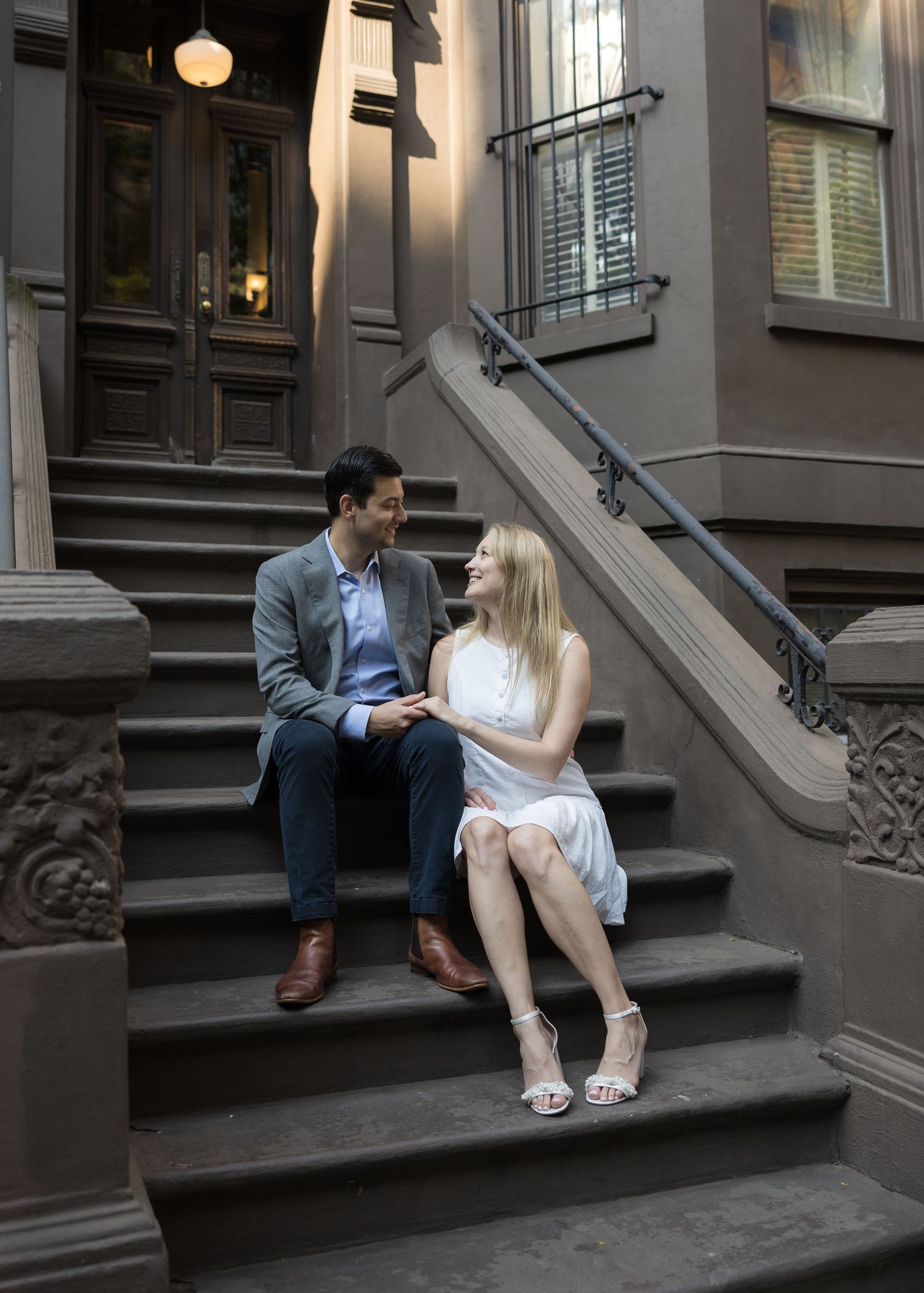 Couple sitting on brownstone steps in NYC, smiling at each other, man in blazer, woman in white dress.