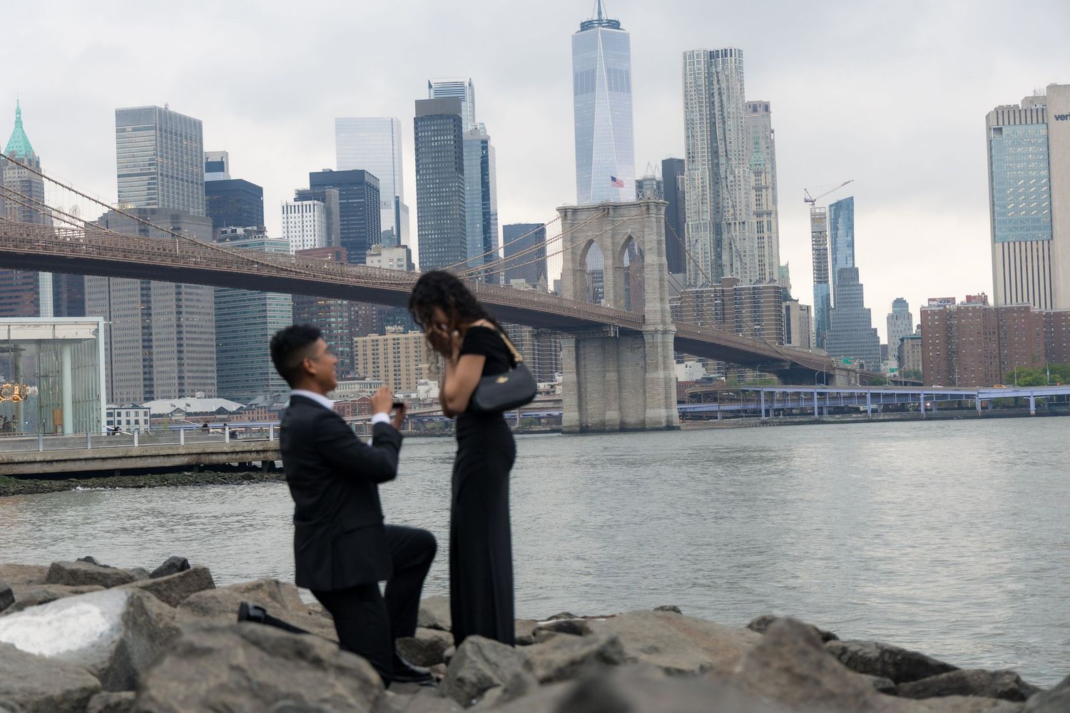 Man proposing to woman on rocky waterfront with NYC skyline and Brooklyn Bridge in background.