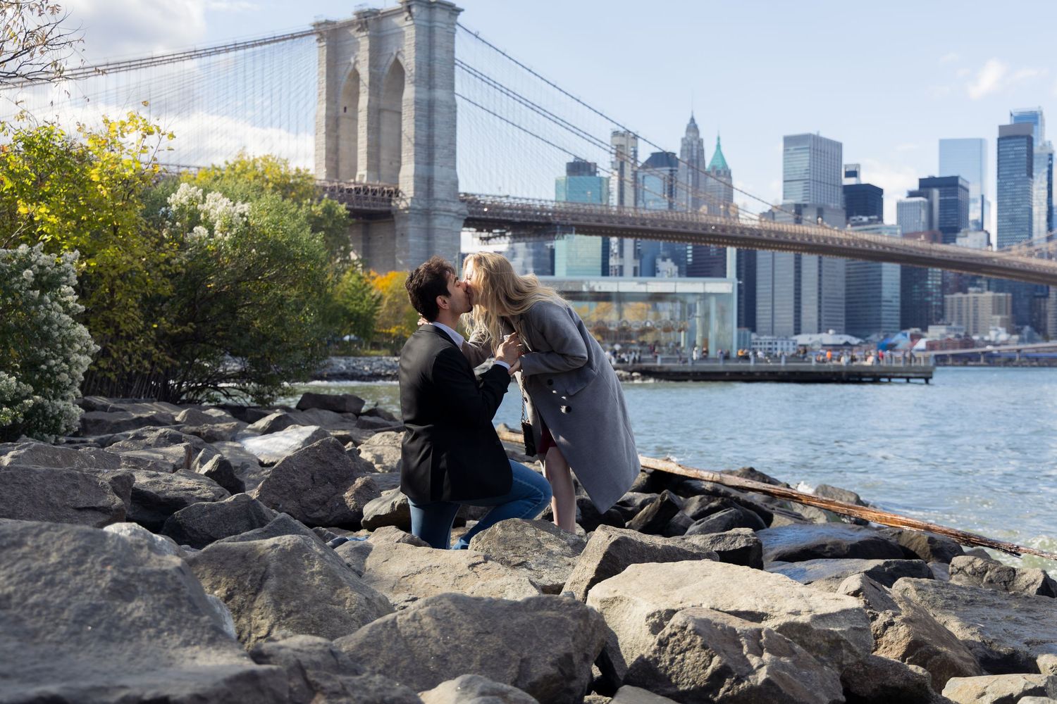 Couple sharing a romantic kiss on rocky shore with Brooklyn Bridge and NYC skyline in the background.