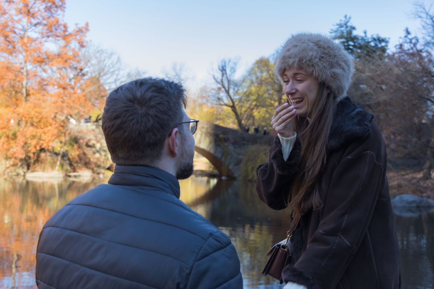 Man proposing to surprised woman by autumn lake in Central Park, she covers mouth in shock.