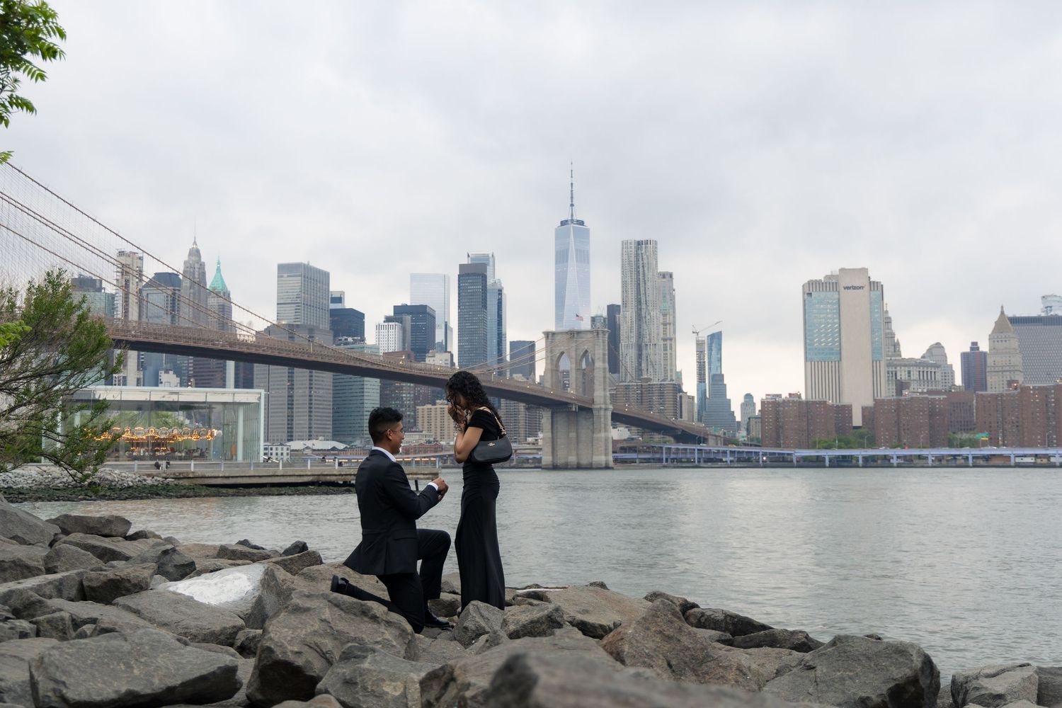 Man proposes to woman on rocky shore with Brooklyn Bridge and NYC skyline in background.