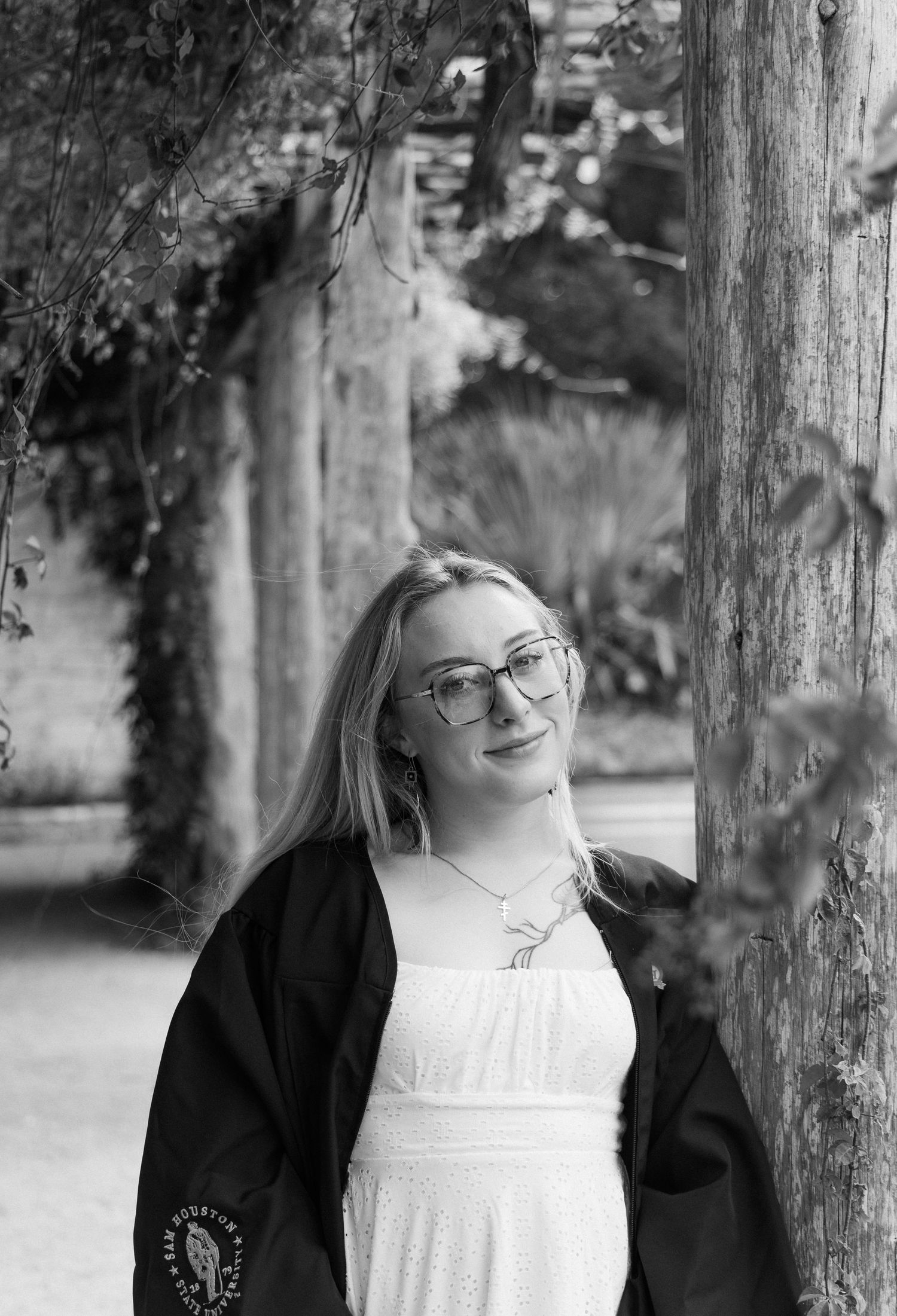 Black and white portrait of a smiling blonde woman with glasses wearing a graduation gown.