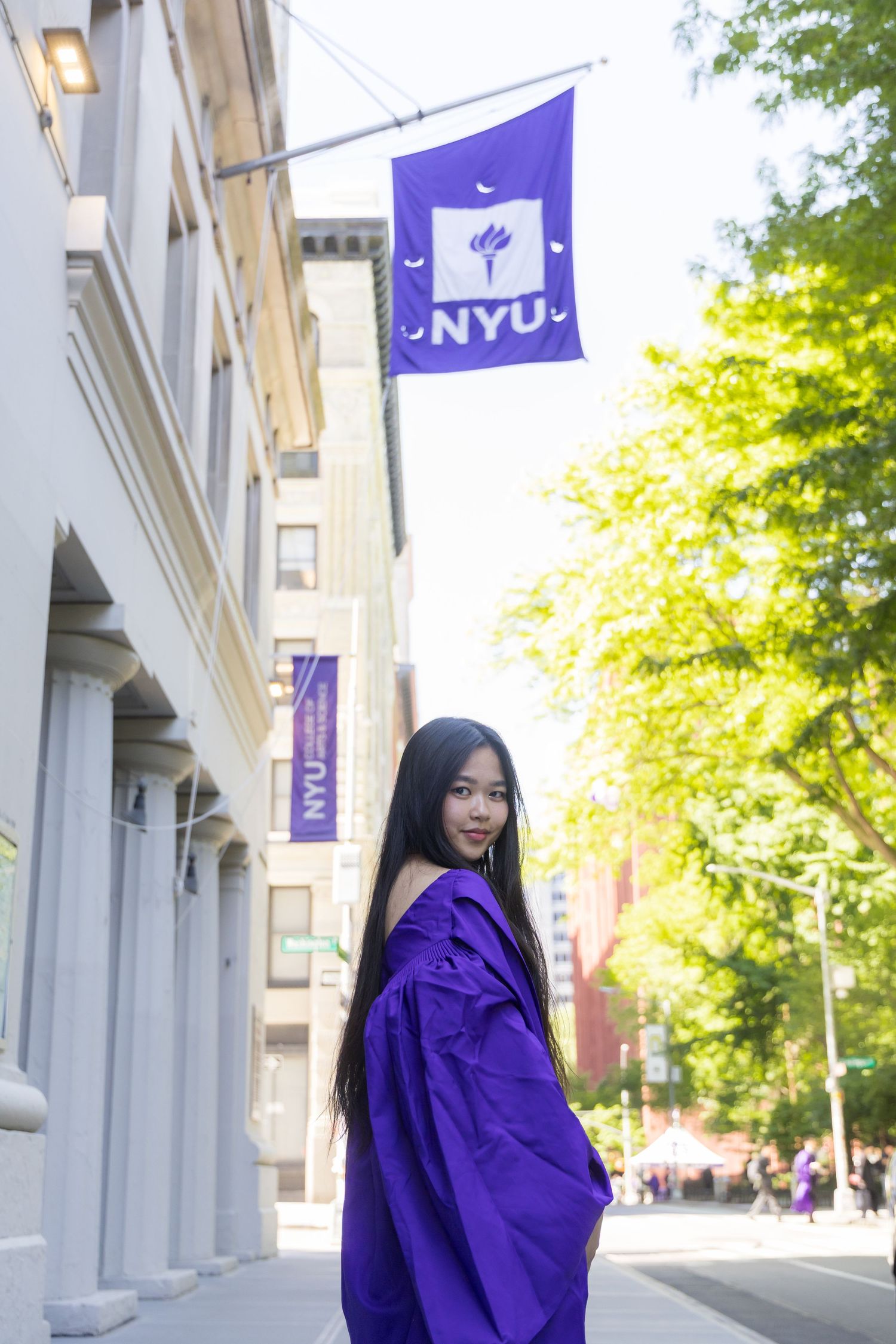 Young woman in purple graduation gown poses on NYC street near NYU banner and flag on sunny day.