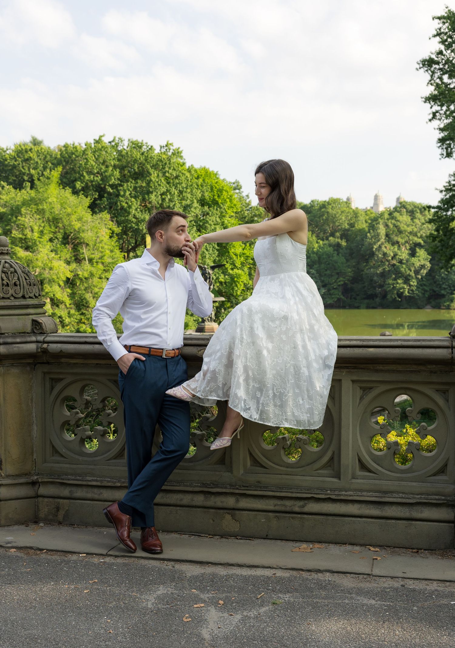 Couple in wedding attire posing on ornate stone bridge in Central Park, woman in white dress sitting on railing.