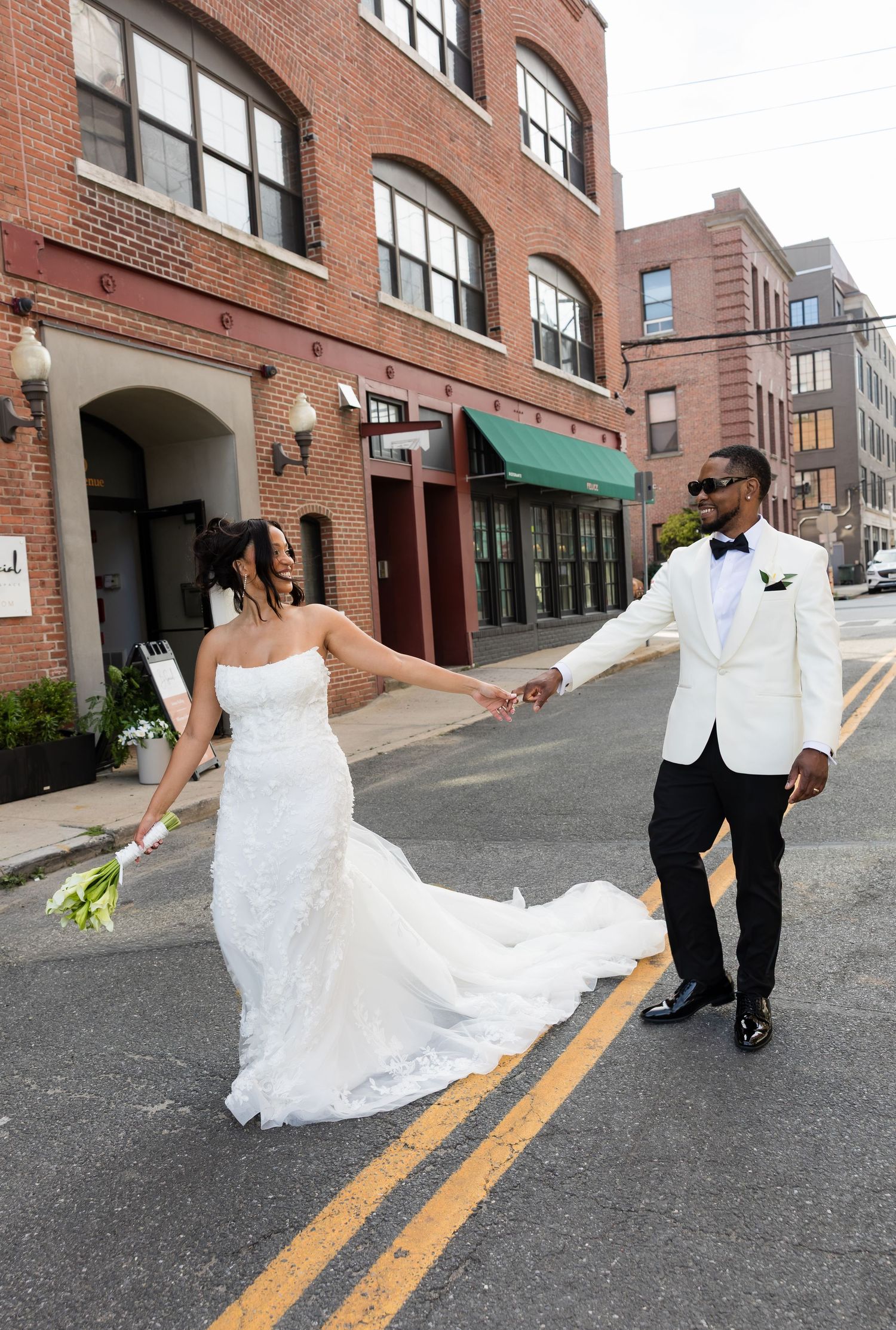 Bride in white gown and groom in white tuxedo hold hands crossing a city street on their wedding day.