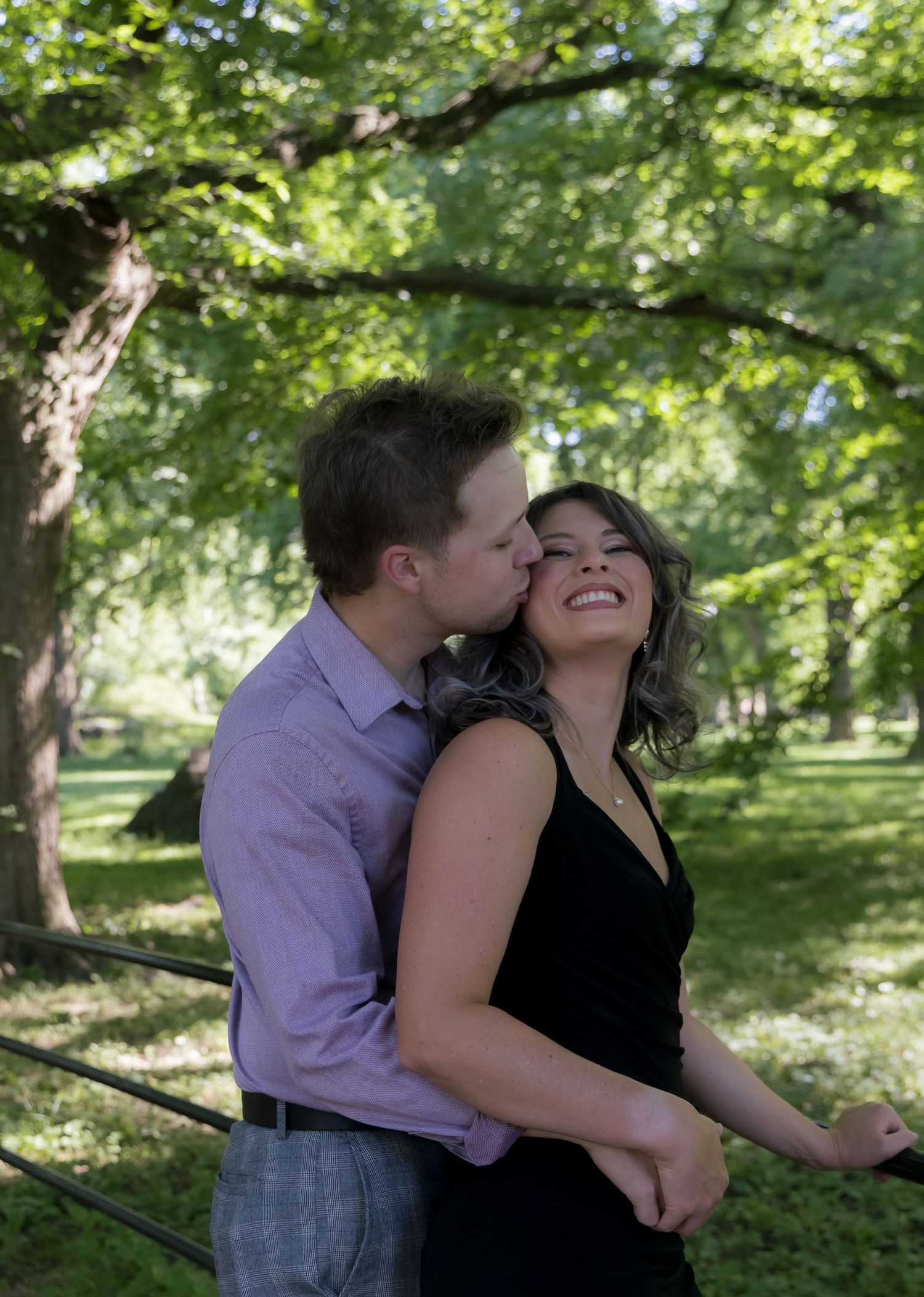 A couple shares a romantic kiss in a lush green park, the woman smiling joyfully in a black dress.