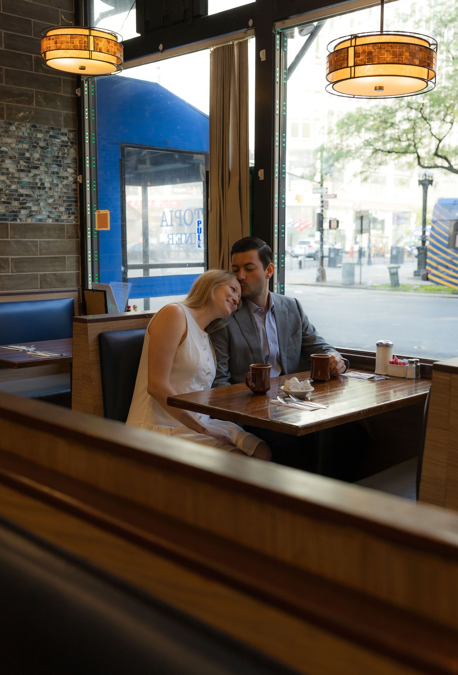 Couple sharing an intimate moment in a cozy restaurant booth near a window, with warm pendant lights above.